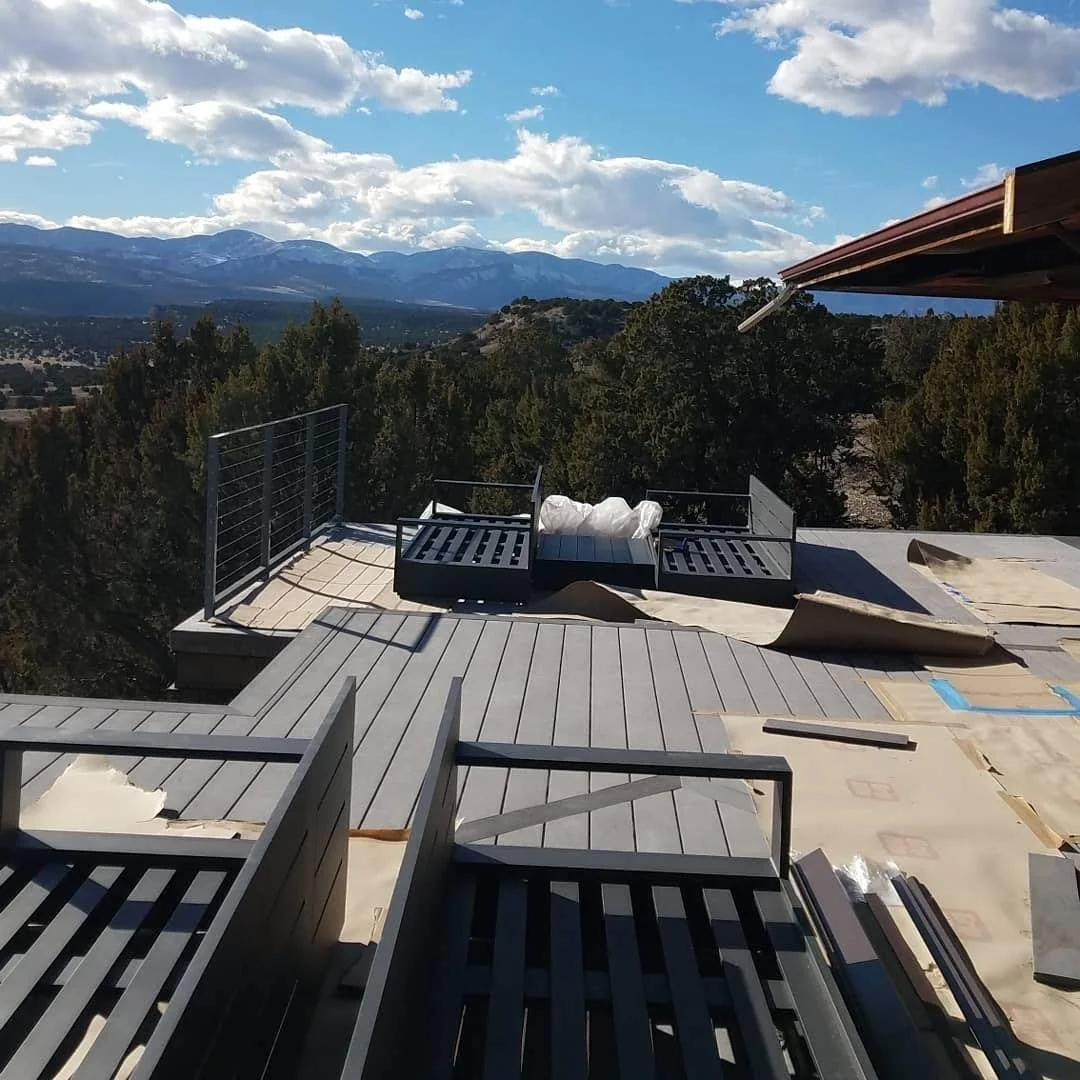 Construction site on a rooftop with partially installed deck and protective barriers, overlooking a mountain range and forest under a partly cloudy sky.