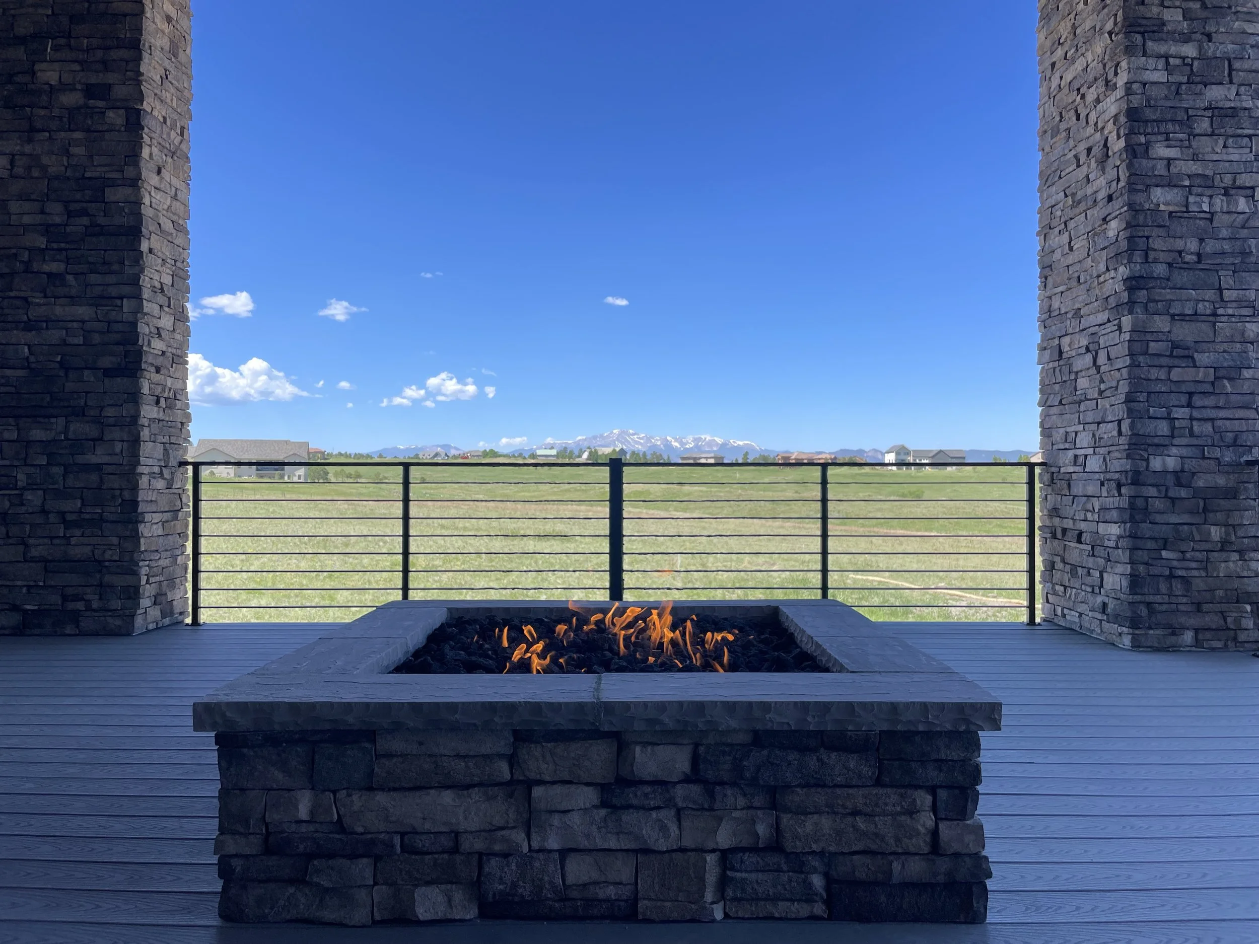 View of a scenic backyard with a Trex deck and square stone fire pit, framed by two stone columns, overlooking a grassy landscape with Colorado Front Range mountains under a blue sky with a few clouds.