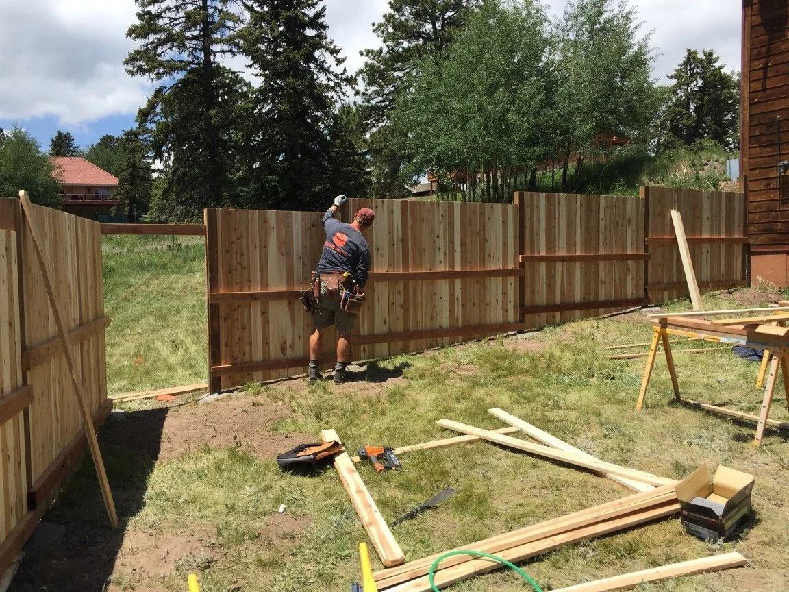 A man installing a wooden fence in a backyard during daytime, with building supplies and tools on the grass.
