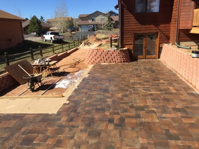 Backyard patio under construction with interlocking bricks, wheelbarrow, and tools.