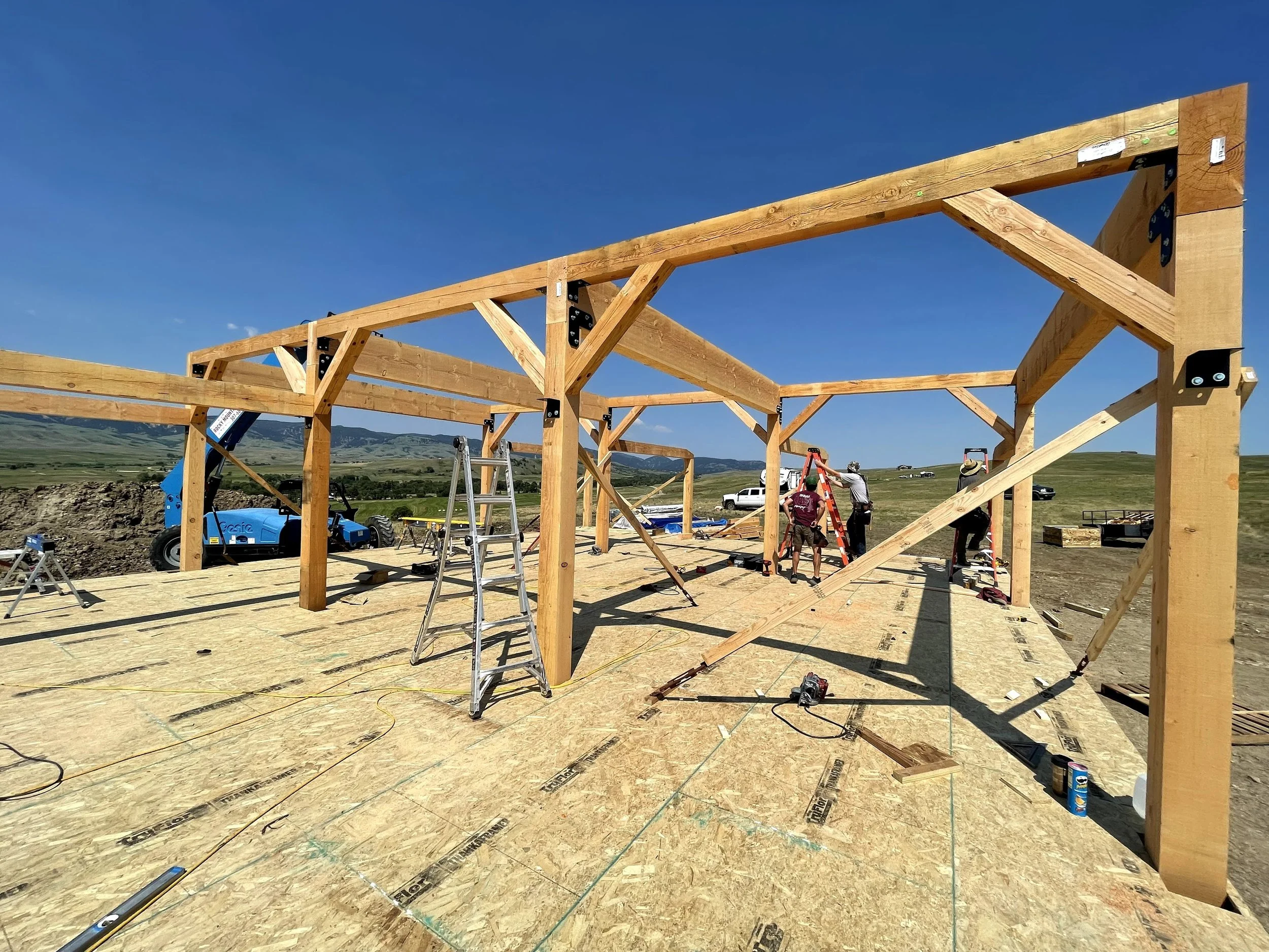 Construction workers building Riverbend Post and Beam home, with ladders, tools, and equipment, under a clear blue sky.