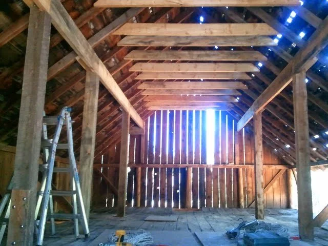 Inside a wooden barn attic with exposed beams and a ladder leaning against the left wall, construction tools on the floor, and light coming through gaps in the wood.
