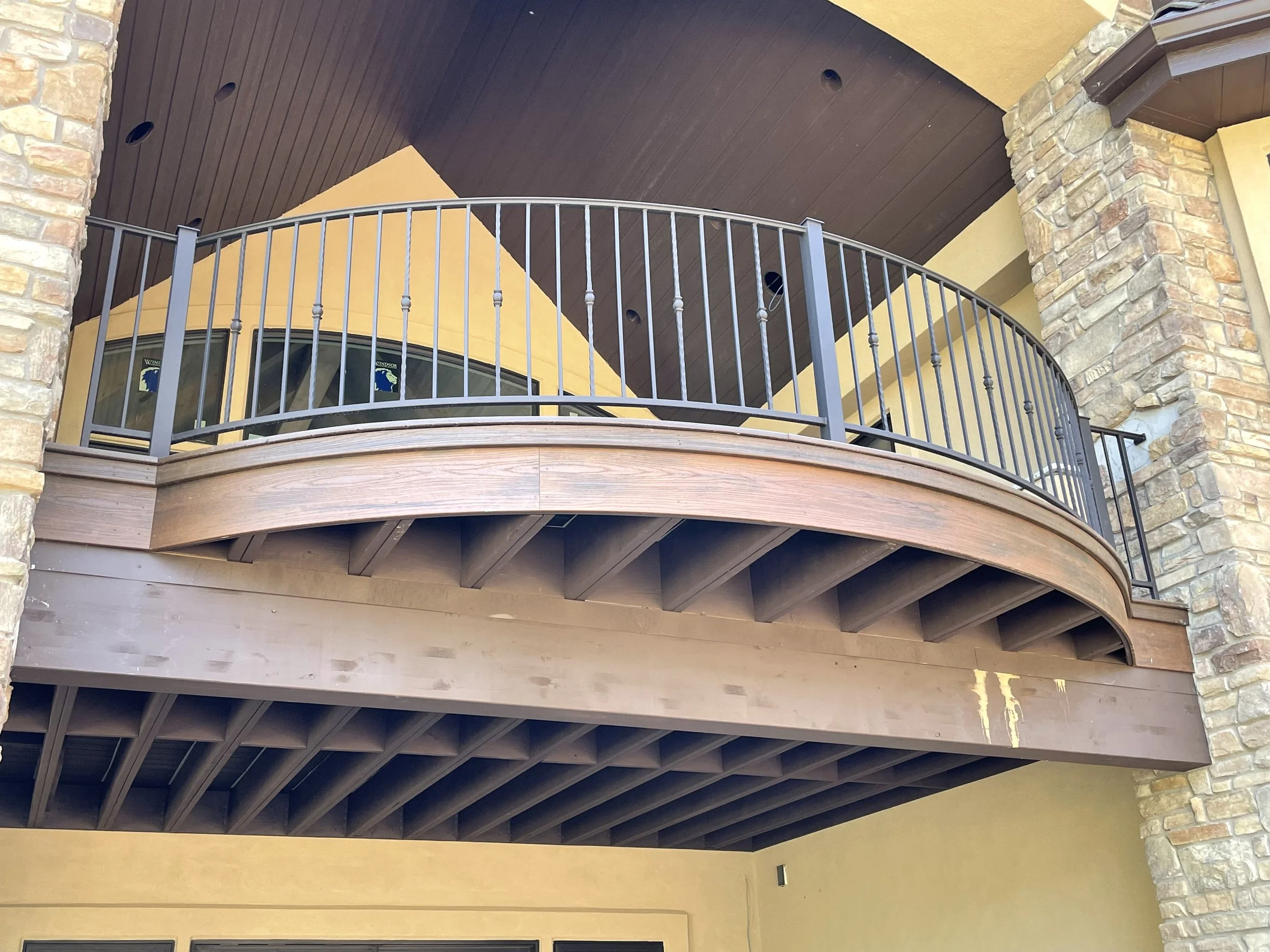 Ceiling of a covered balcony with a dark brown wood finish, surrounded by a metal railing and stone columns supporting the structure.