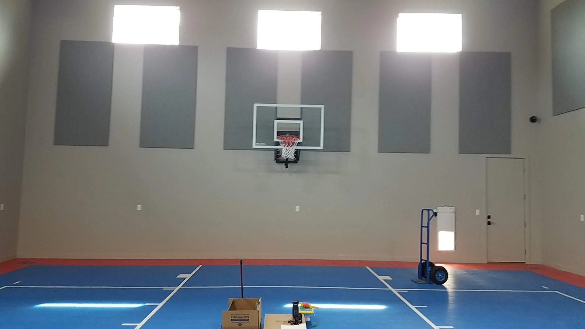 An indoor basketball court with gray acoustic panels on the wall, three high windows, a basketball hoop, a blue cart, and some construction tools and materials on the floor.