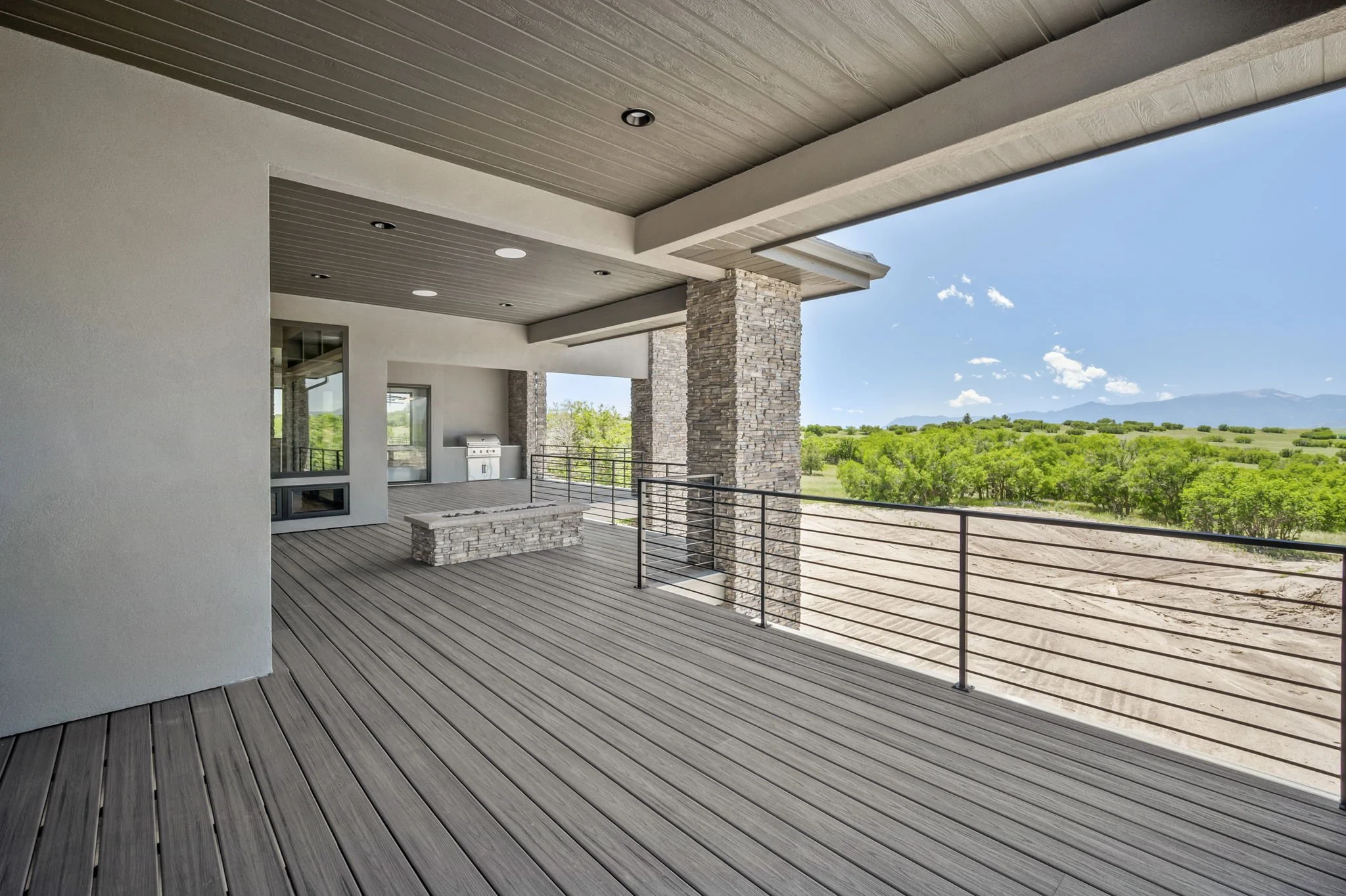 Modern house balcony with wooden flooring, stone pillars, and a view of a green landscape with distant mountains under a blue sky.