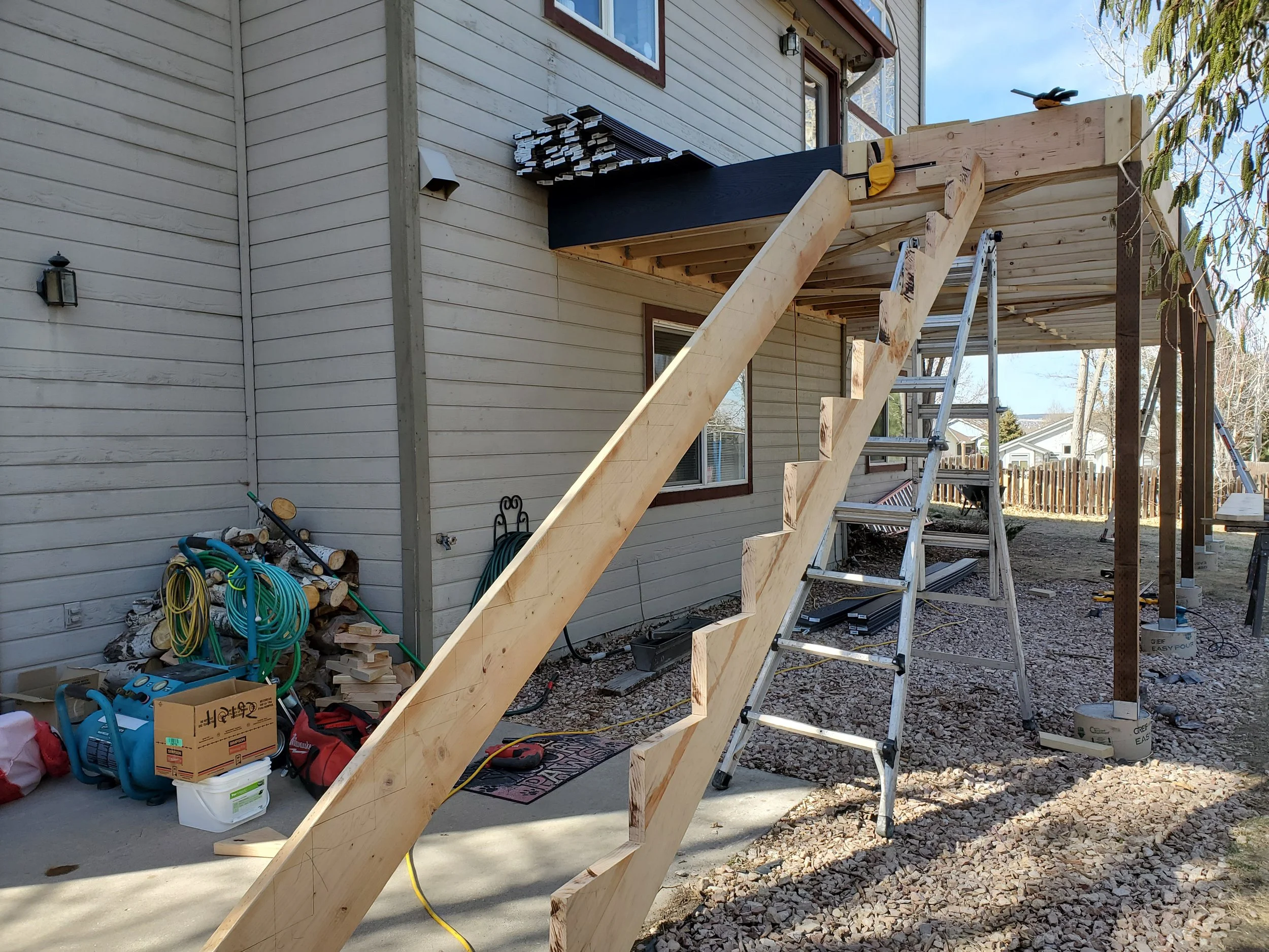 Construction site with wooden staircase in progress outside a house. Various tools, building materials, and equipment scattered around, including hoses, a ladder, and boxes.