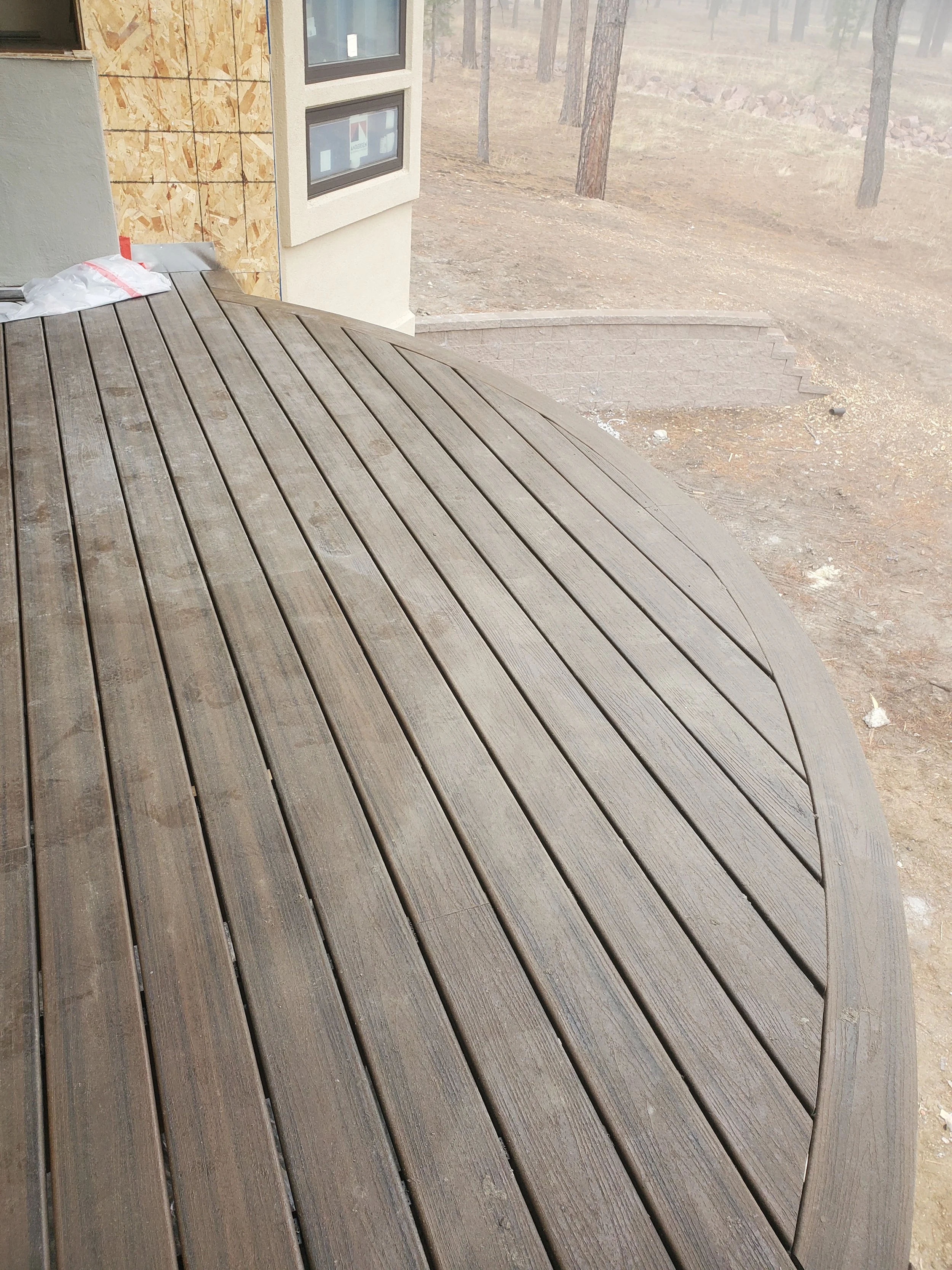 A wooden deck with parallel planks outside a house under construction, with a semi-wooden wall and forested area in the background.