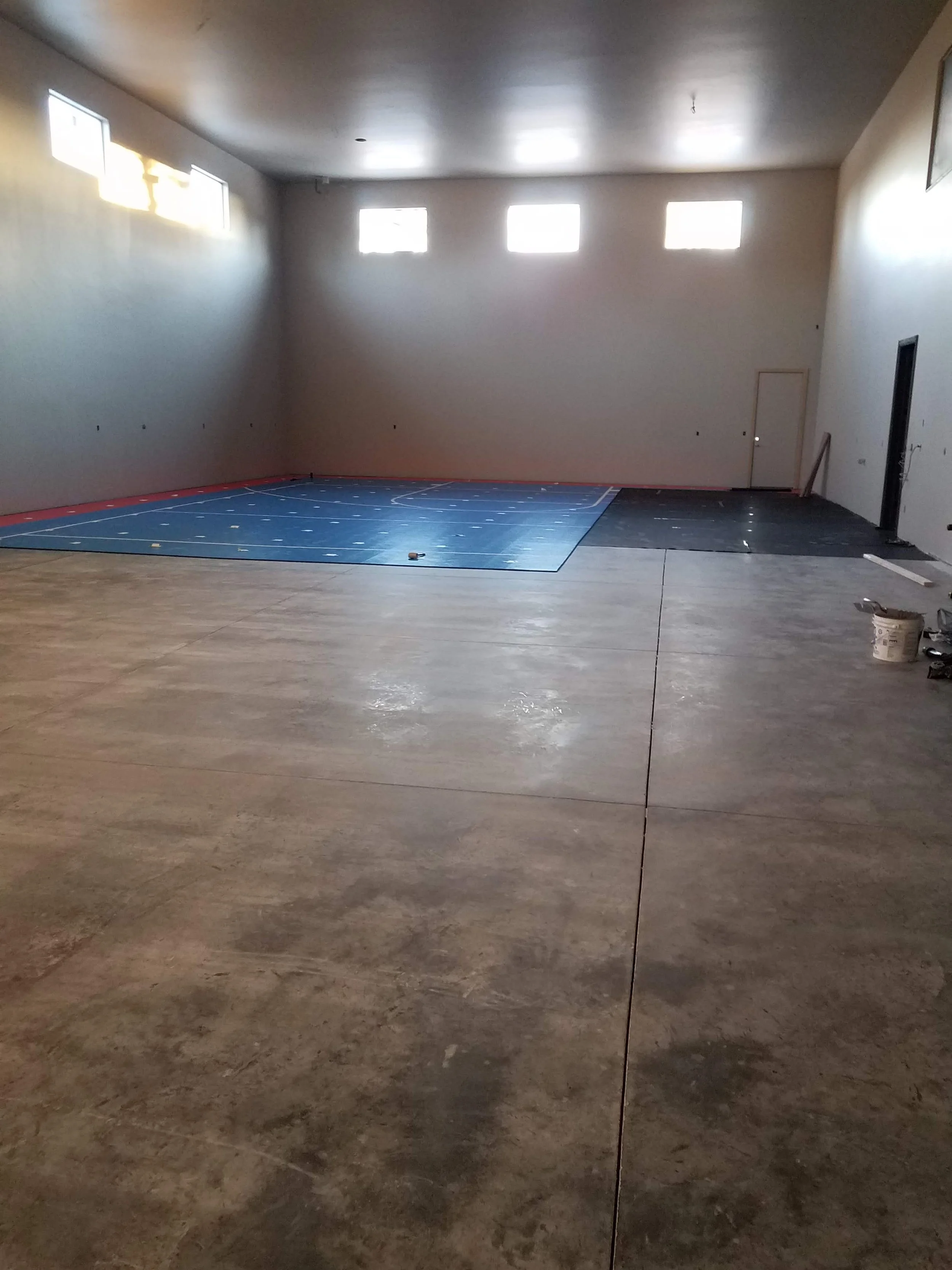 Interior of a room under construction with a basketball court floor, unfinished walls, and windows near the ceiling.