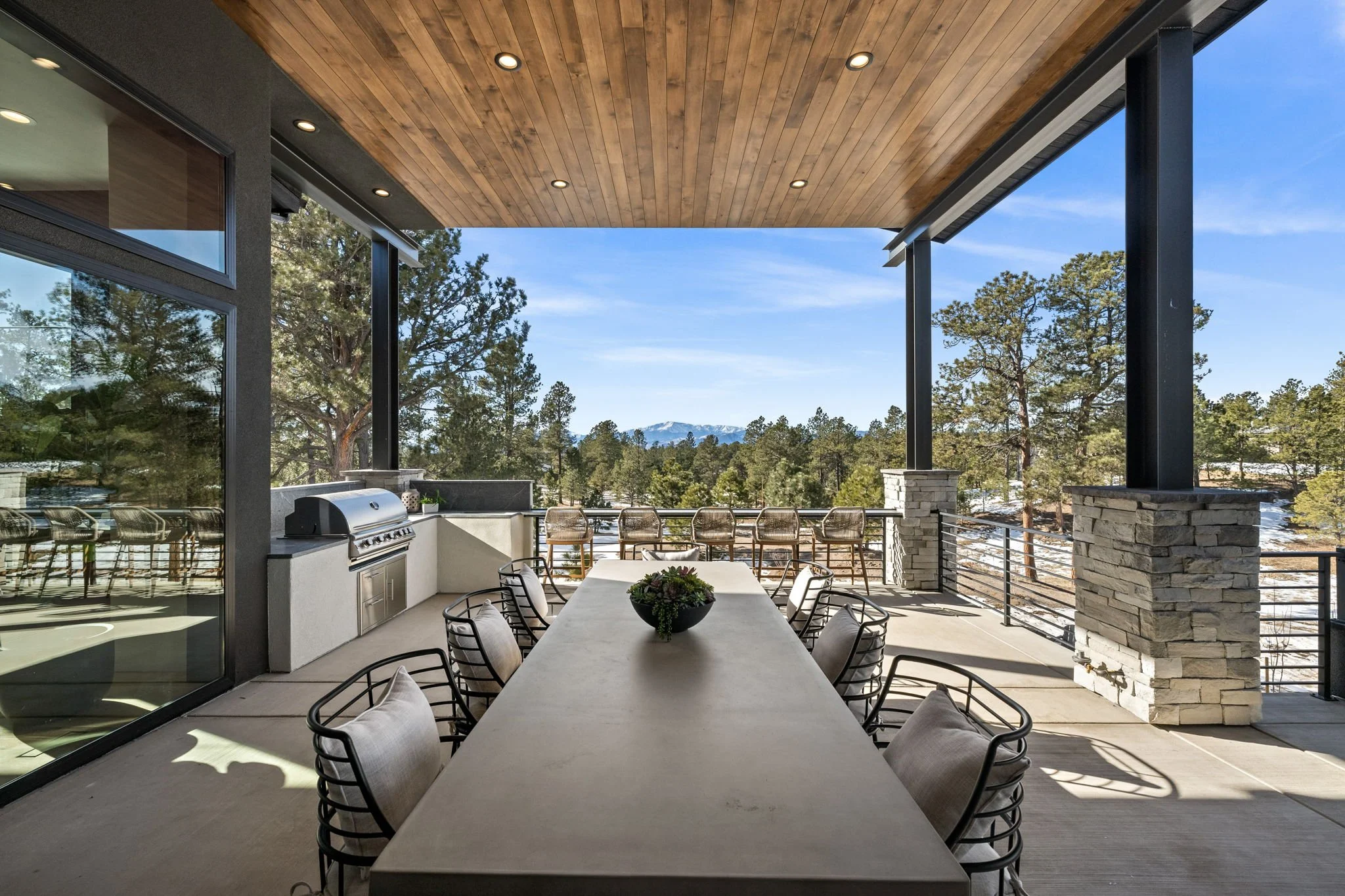Outdoor balcony or patio with a long dining table, chairs, and built-in barbecue grill, overlooking a forested landscape with distant mountains and a blue sky.