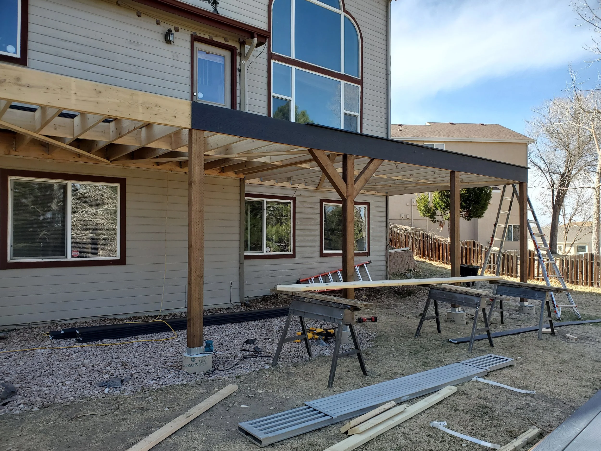 Construction site with a house in the process of having a deck built. There are wooden beams, support posts, construction tools, and materials scattered around the area.