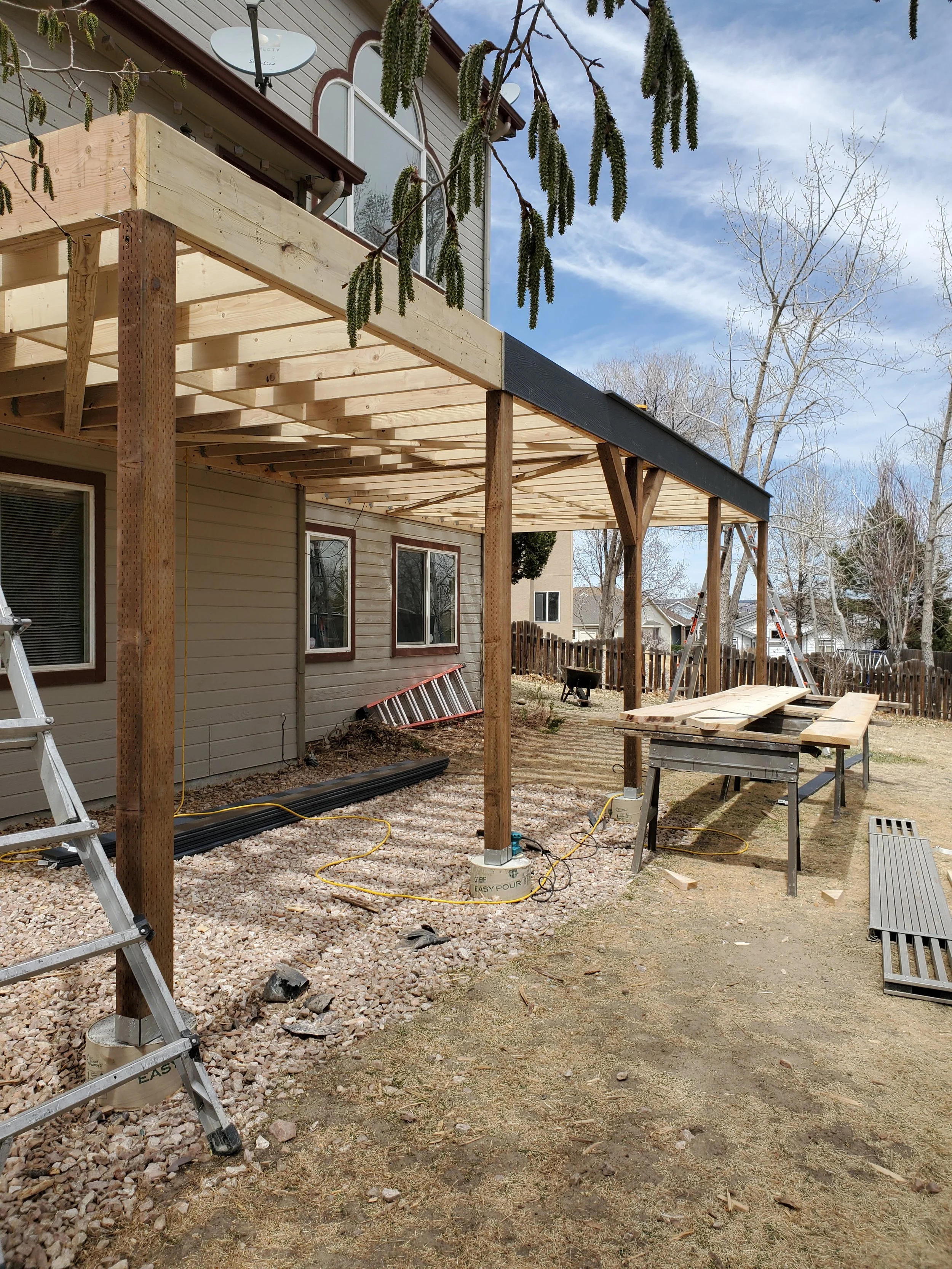 Construction site with an unfinished deck extension on the back of a house. Wooden beams and posts are in place, with some tools and construction materials around, including ladders, a work table, and a wheelbarrow. The area is partially covered with