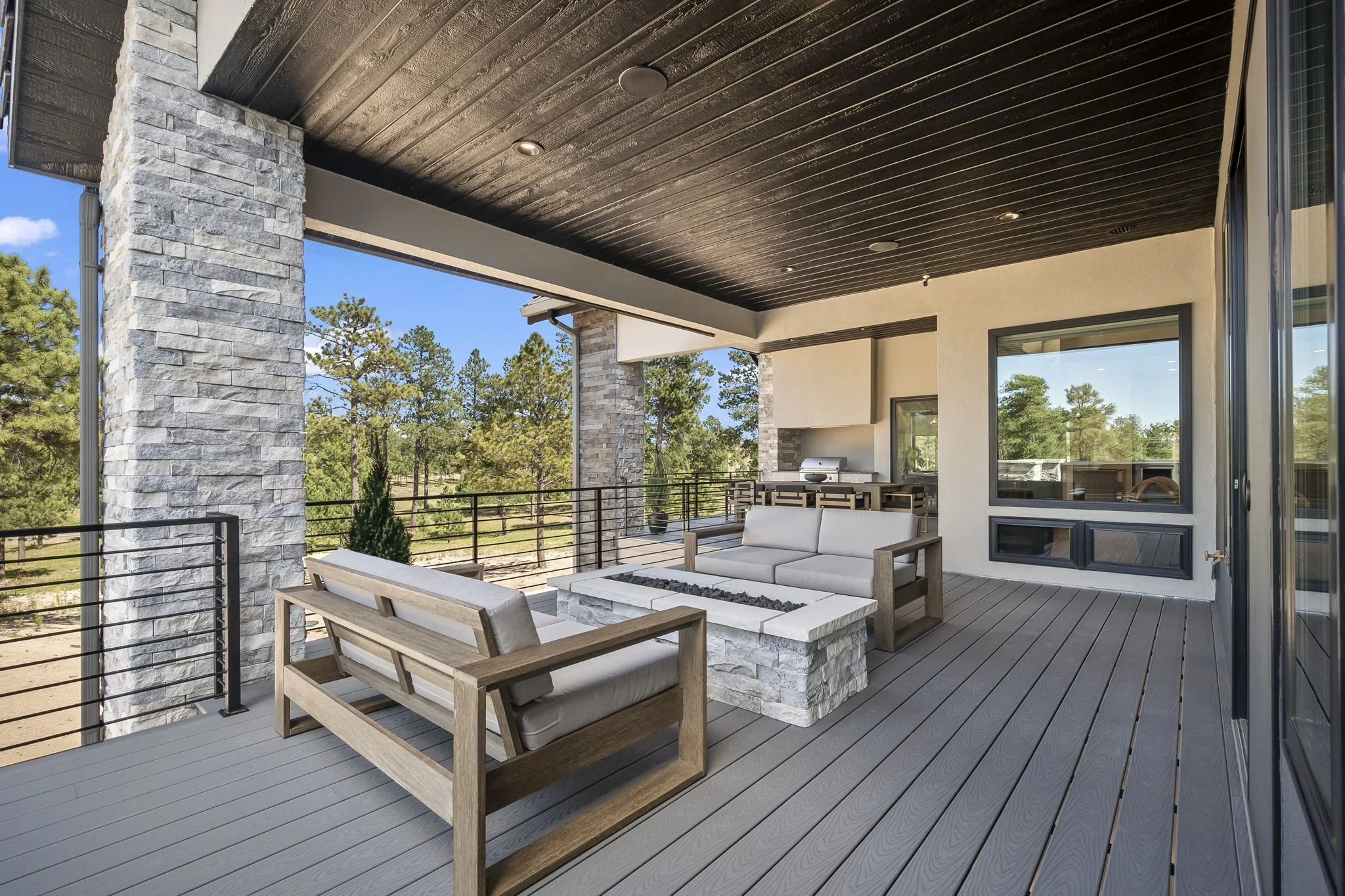 A modern outdoor balcony with a dark wood ceiling, stone pillars, and a railing. The balcony has outdoor seating including wooden furniture with white cushions around a firepit, and a built-in grill area. Trees and blue sky are visible in the backgro