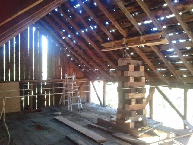 Interior of a historic barn under renovation with exposed wooden beams and a cribbing stack with a pump jack to raise the roof, construction tools, and a ladder.
