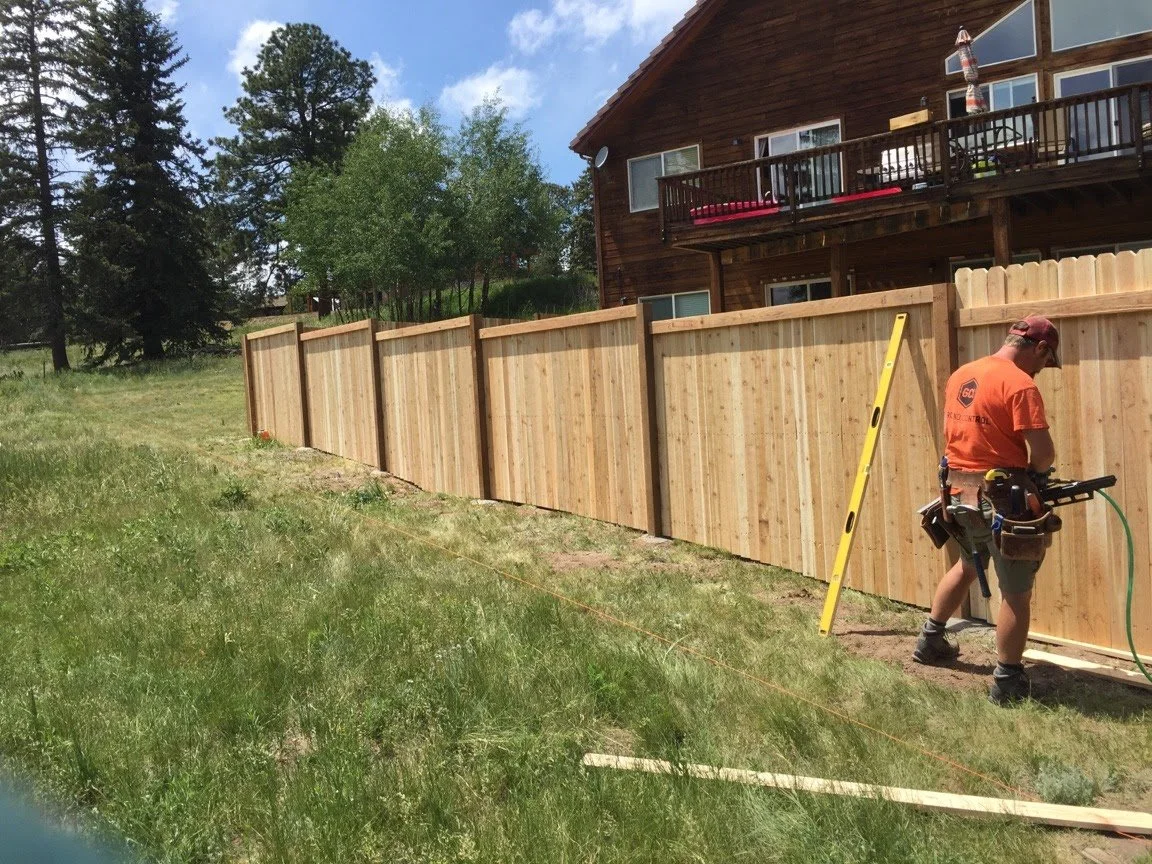 A worker using tools on a wooden fence in a backyard with a house that has a balcony and potted plants.