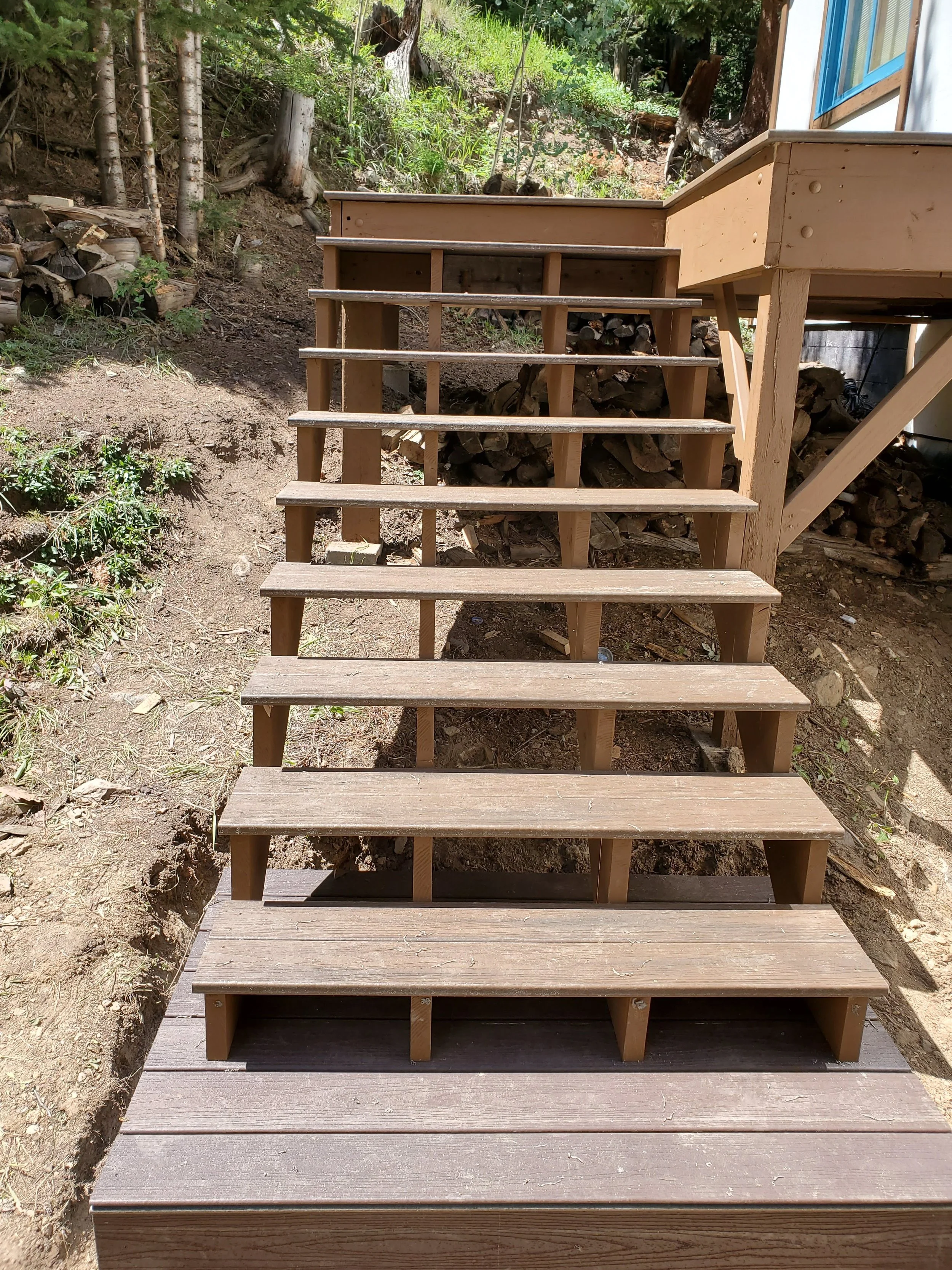Wooden stairs leading up to a raised deck or porch outside a house, surrounded by natural terrain and trees.