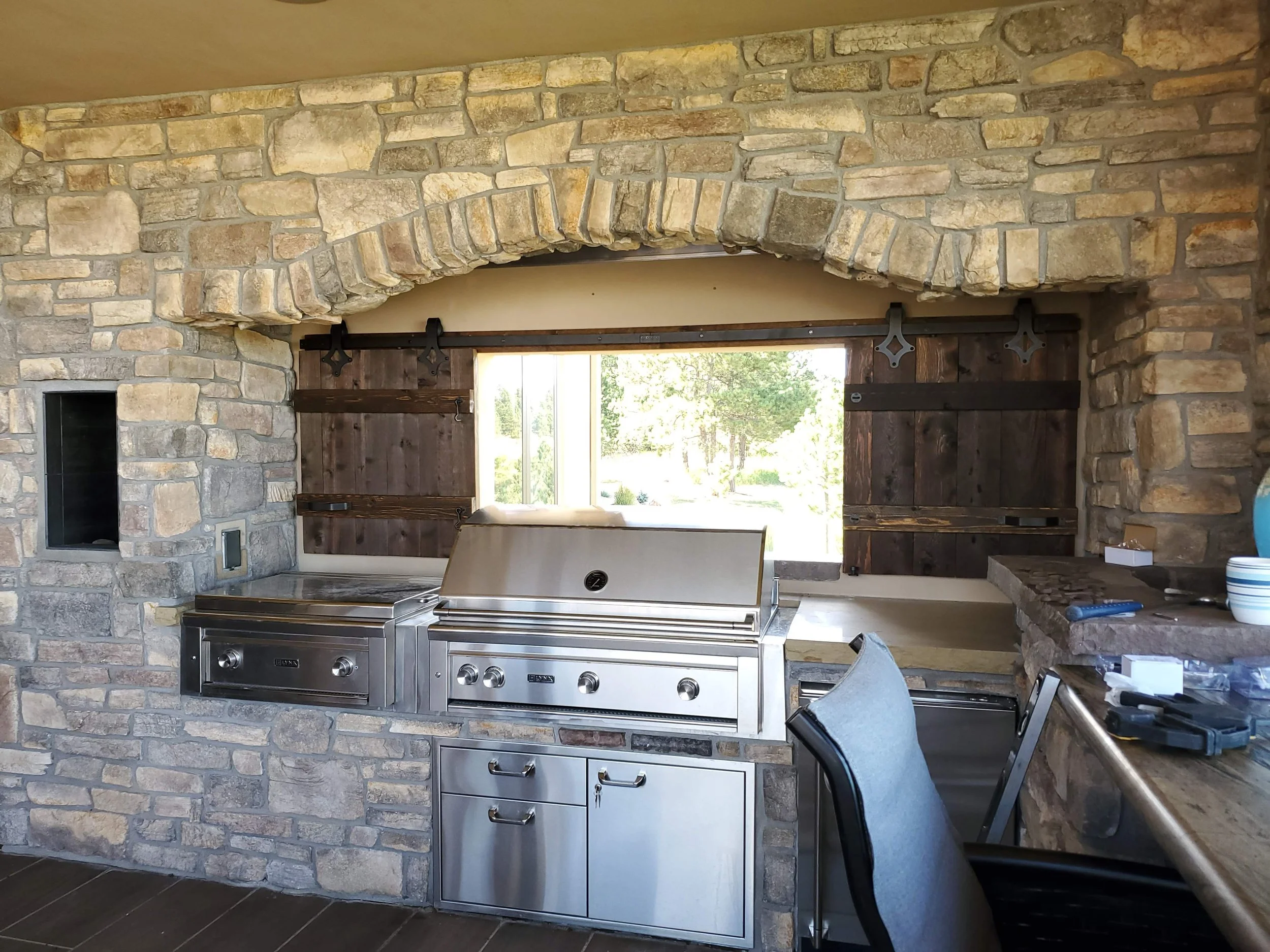 Outdoor kitchen area with stone wall, sliding barn door, stainless steel grill, and window showing greenery outside.
