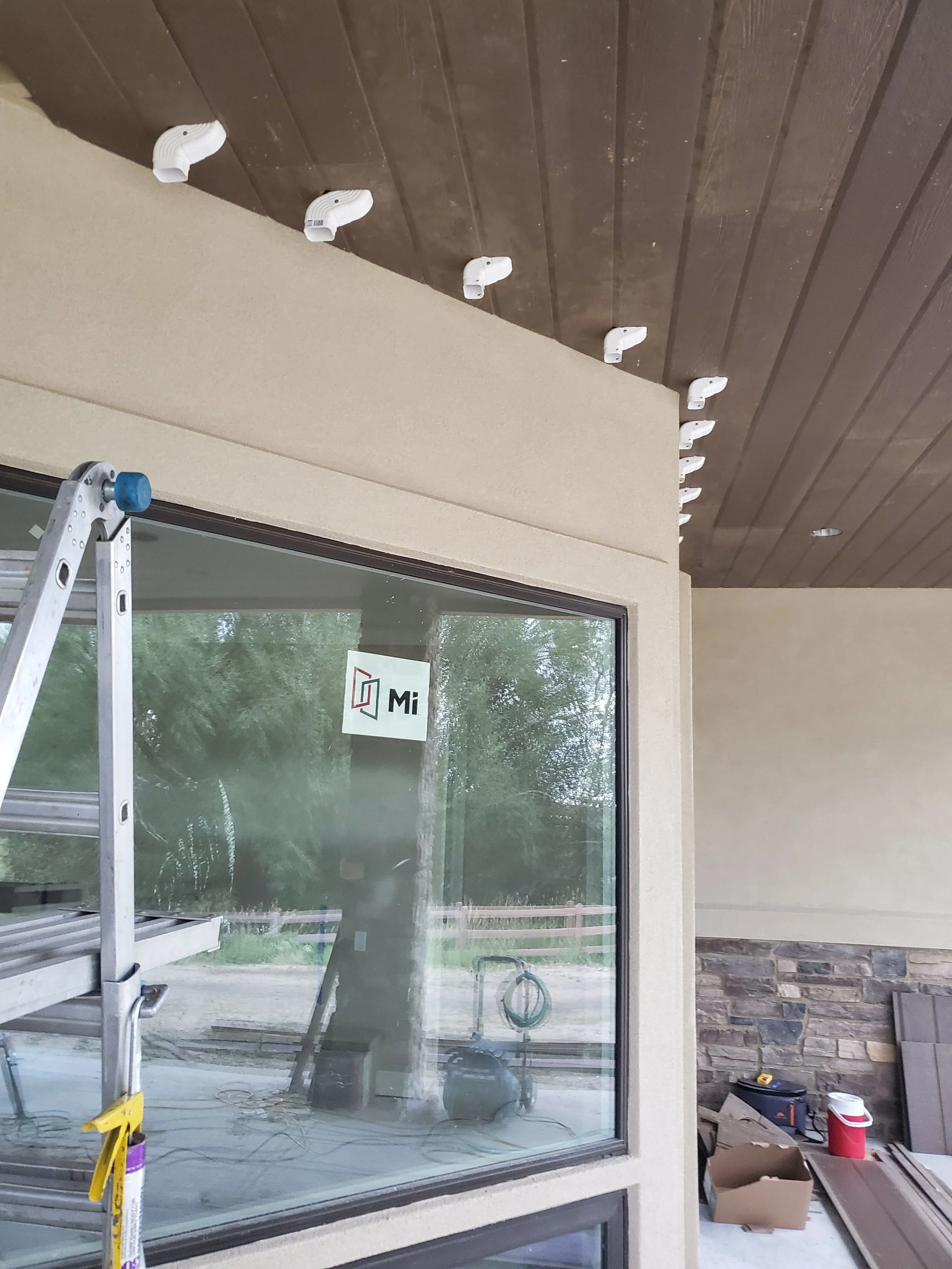 Construction site with a large window glass and a ladder, construction tools, and materials around. The ceiling has wooden panels with multiple white vent covers installed.