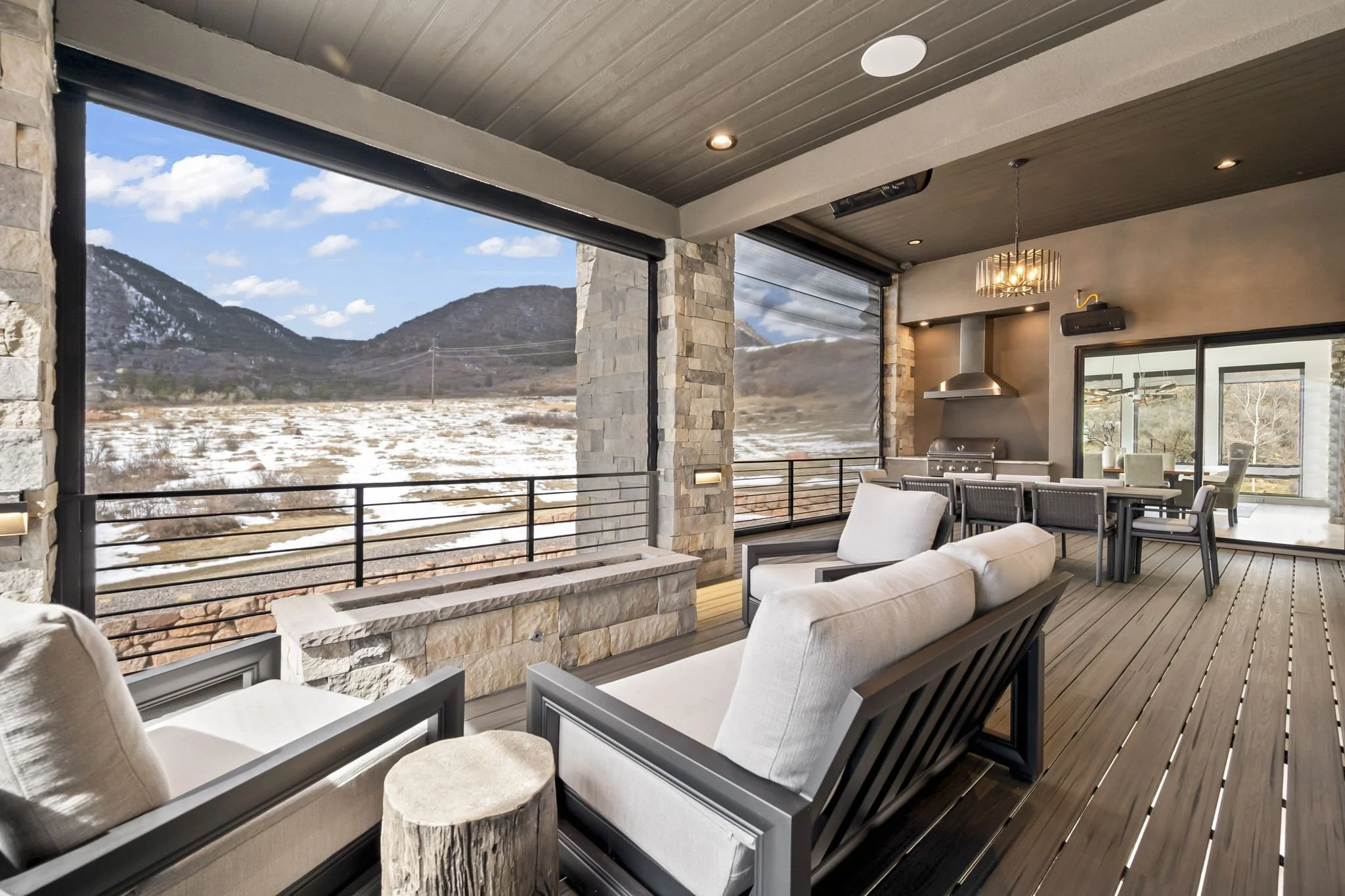 Modern outdoor patio with stone pillars, wooden ceiling, and mountain view; includes seating area with white cushioned chairs and a dining table with chairs, overlooking a snowy landscape and blue sky.