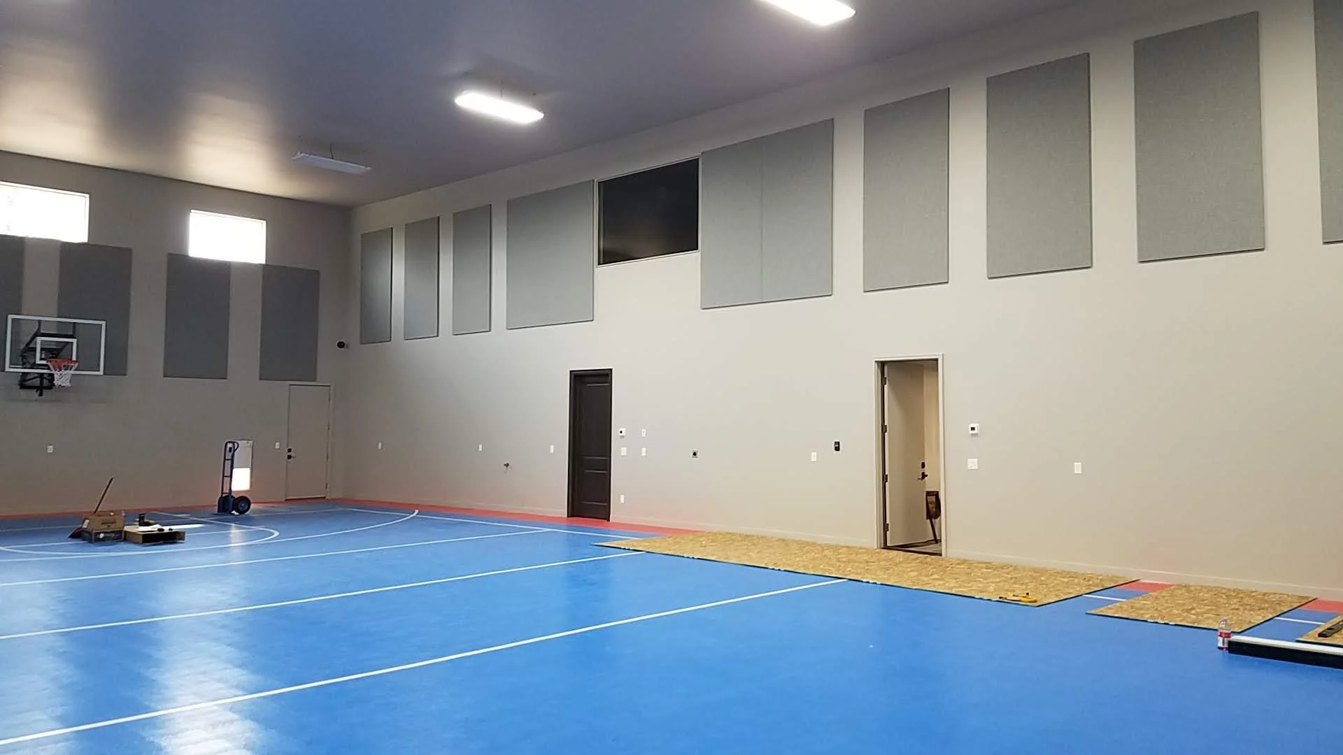 Indoor gymnasium with blue basketball court flooring, basketball hoop on the wall, and soundproof panels on the walls. Some construction or maintenance equipment is on the floor.