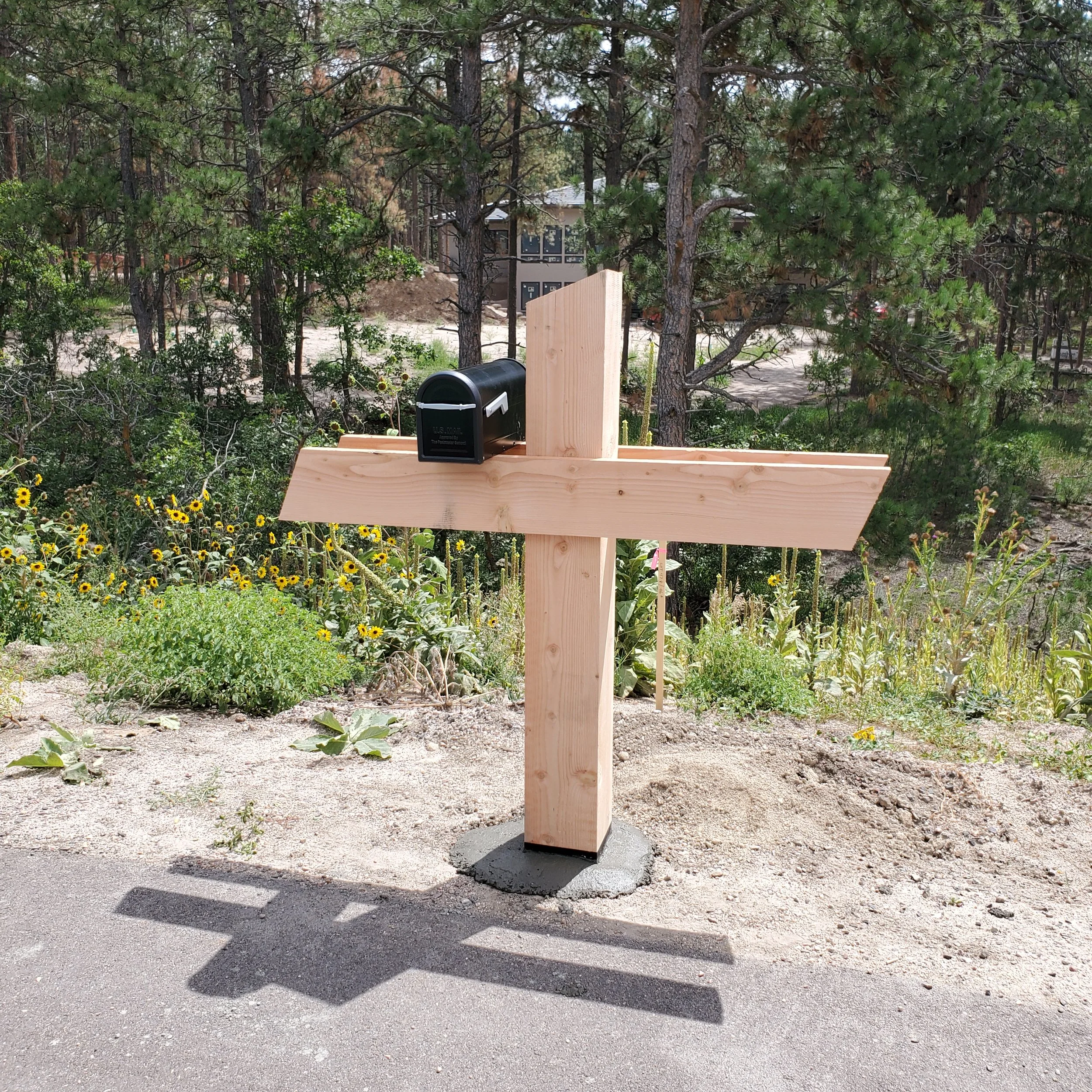 A wooden post next to a black mailbox on a concrete platform, with a background of trees, under a sunny sky casting shadows on the ground.