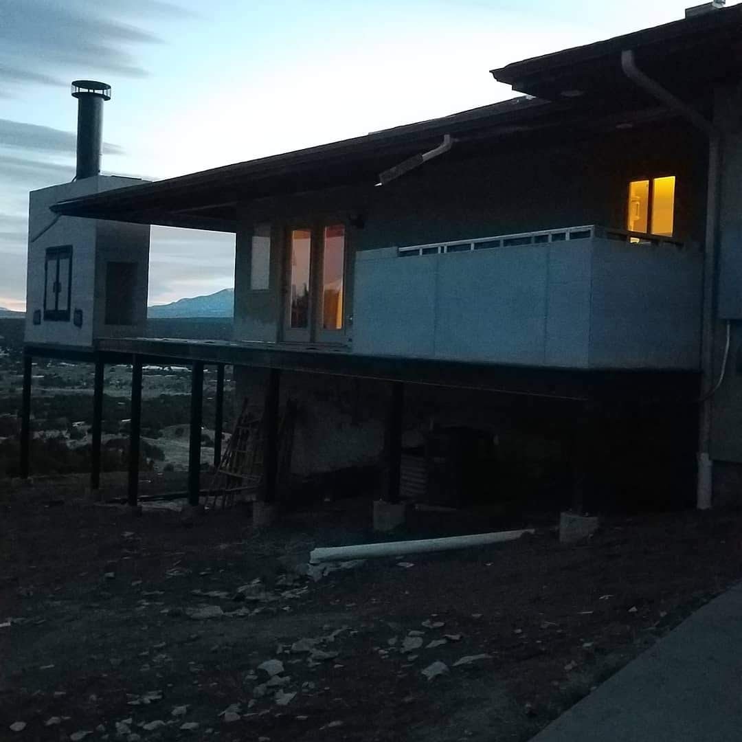 A house elevated on stilts with a dark exterior, windows with warm yellow lights, and a chimney on the roof, set against a dusk sky.