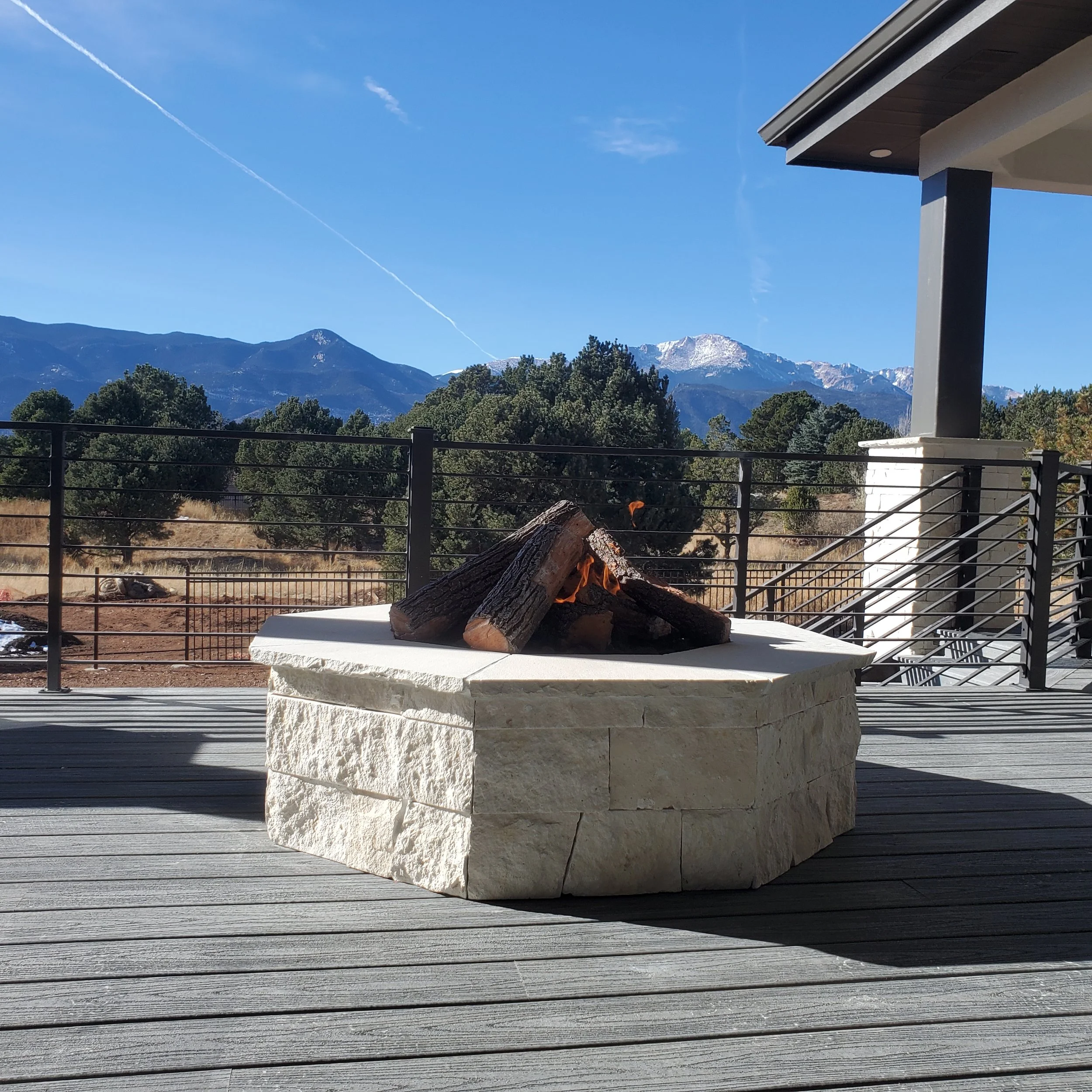 A backyard fire pit with logs burning on a white stone base, set on a wooden deck with a view of mountains and a partly cloudy blue sky.