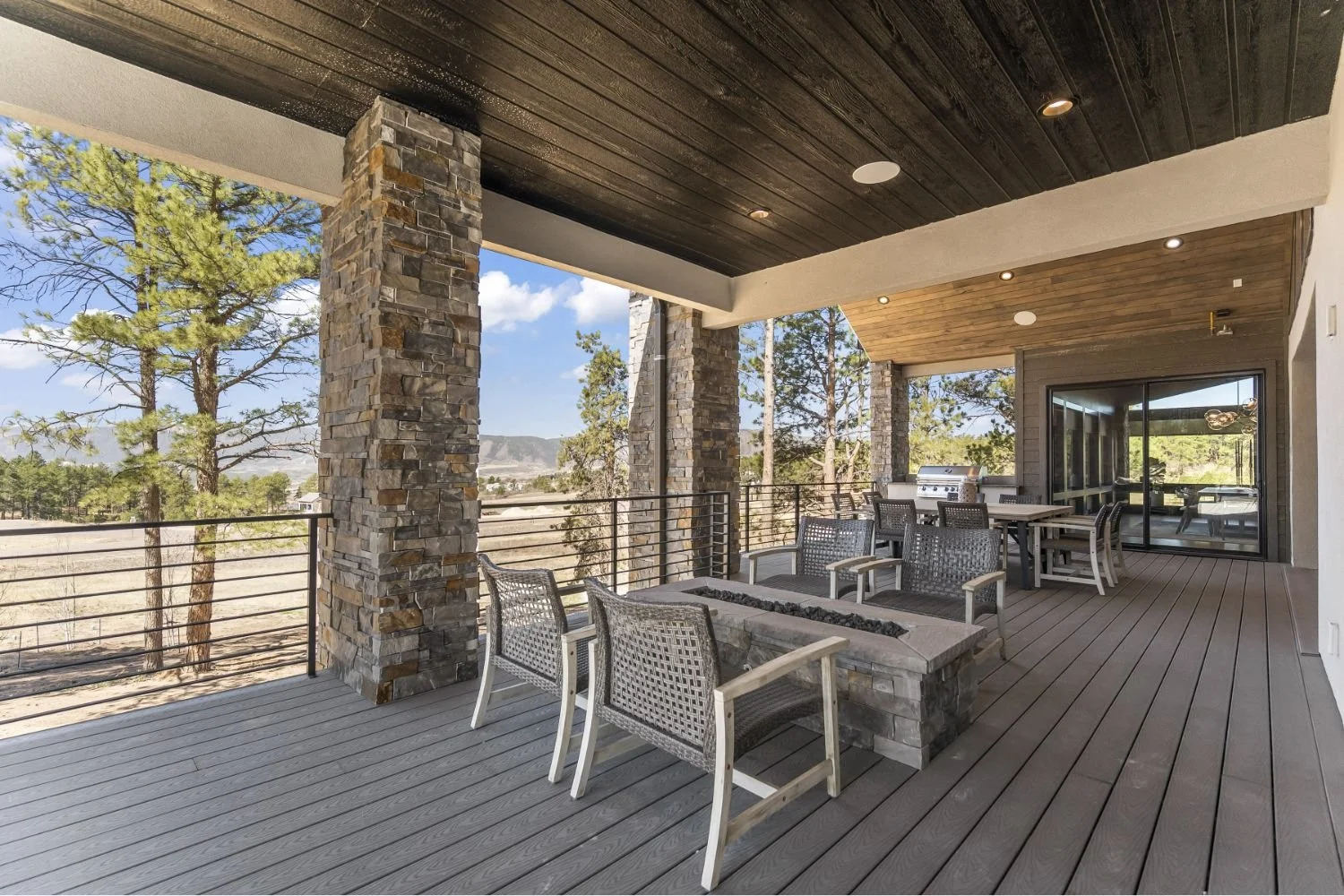 Covered outdoor patio with stone columns, wooden ceiling, and modern furniture, overlooking trees and mountains.