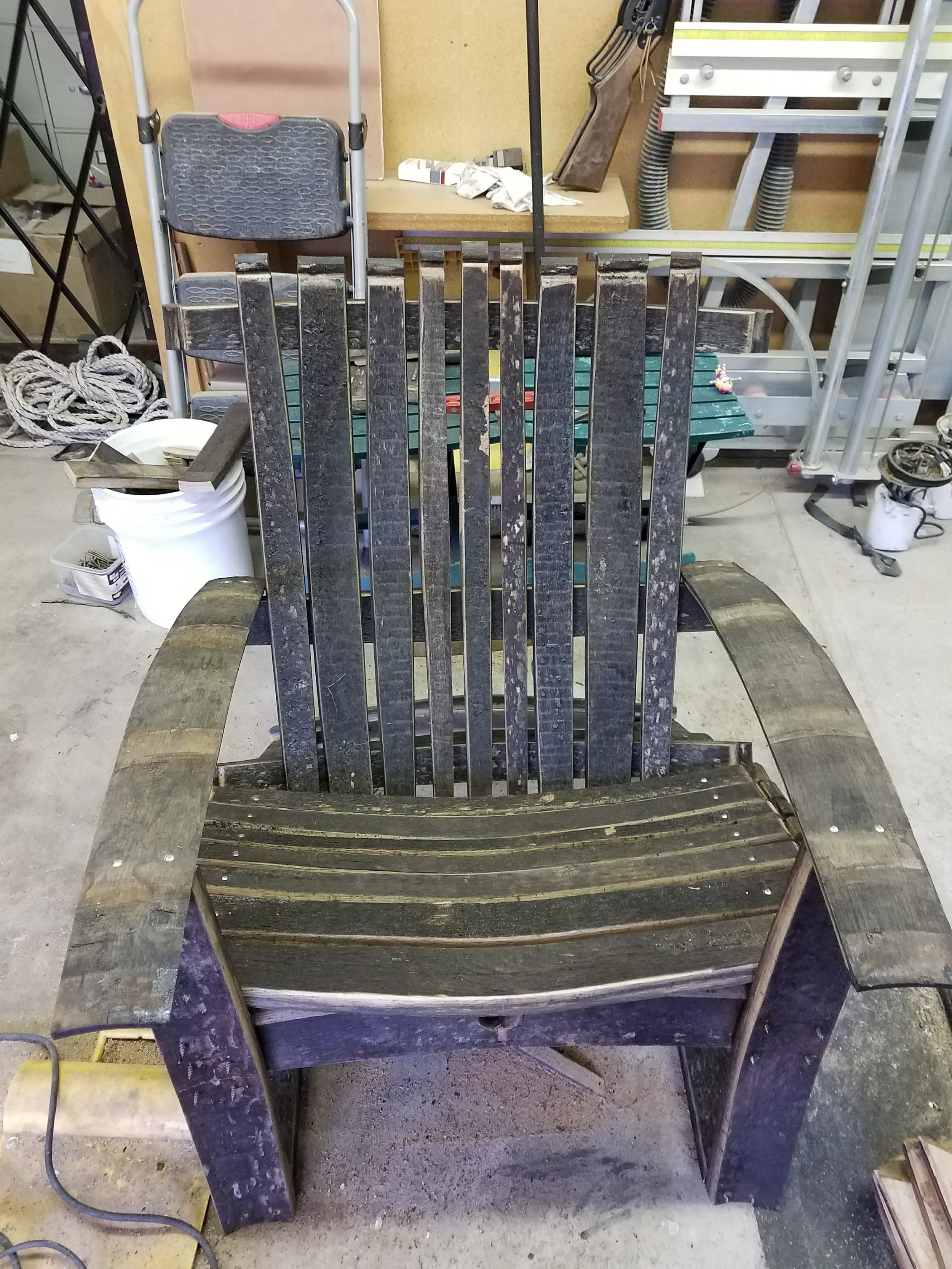 A black wooden Adirondack chair in a workshop or garage, surrounded by tools, a bucket, and hardware fixtures.