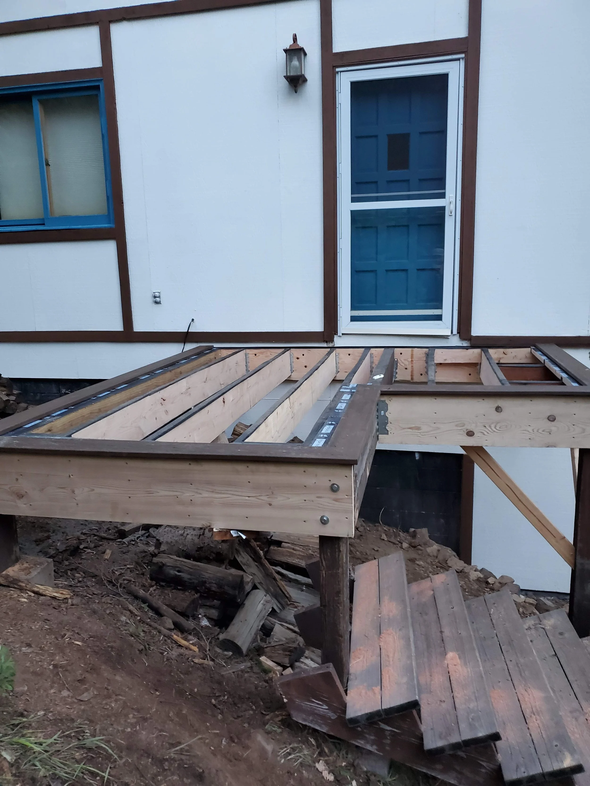 A partially built deck with wooden framing in front of a house with a door and a window. The house has white siding with brown trim, and an outdoor light fixture is mounted above the door. The ground beneath the deck is dirt with some debris.
