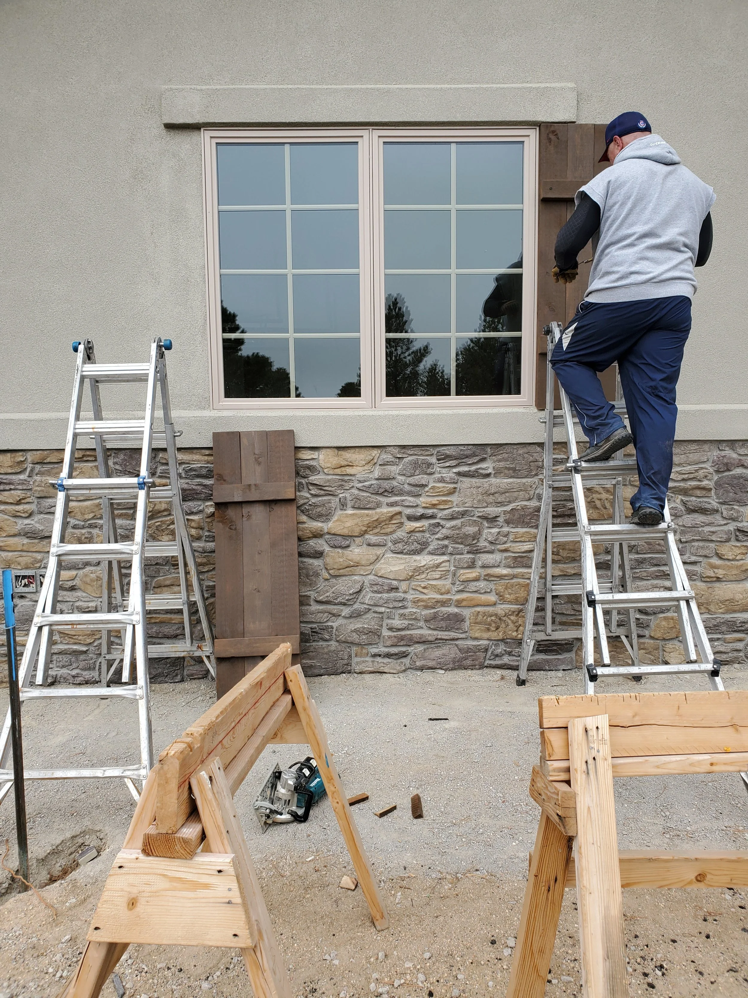 A person working on installation of Cedar shutters on the outside a house, using a step ladder, with construction tools and wood around.