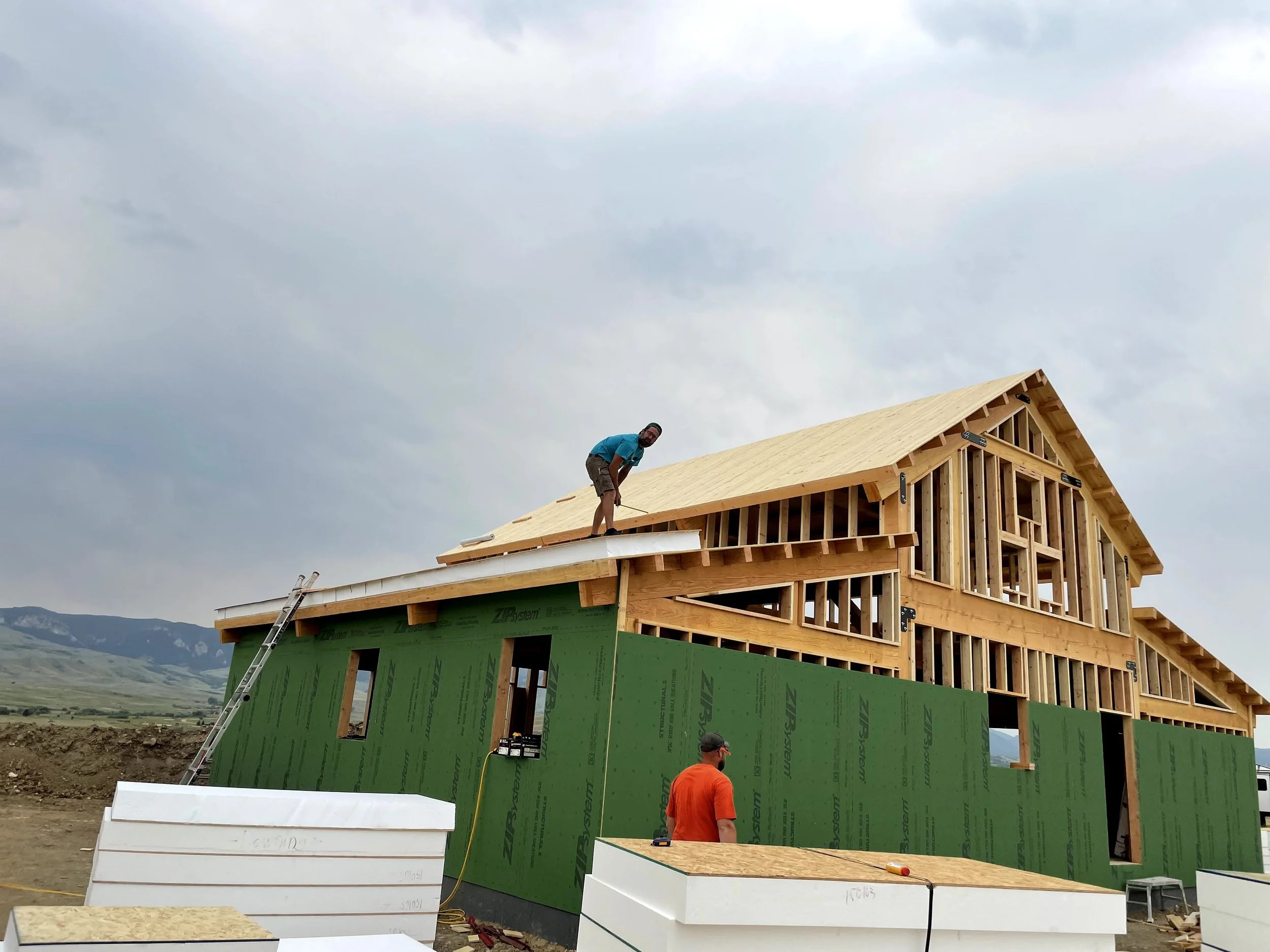 A man working on the roof of a partially constructed house, with Zip sheathing and a wooden frame, against a cloudy sky and mountainous landscape.