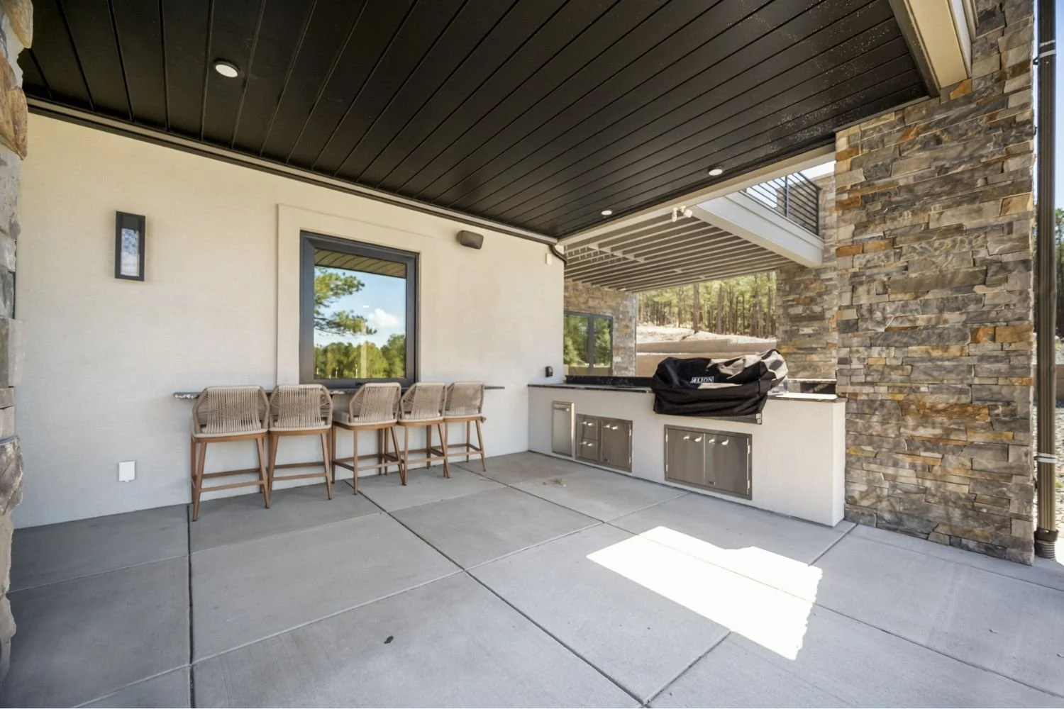 Outdoor covered patio area with concrete flooring, stone wall accents, bar-height chairs, and an outdoor kitchen with a grill covered by a black cover.