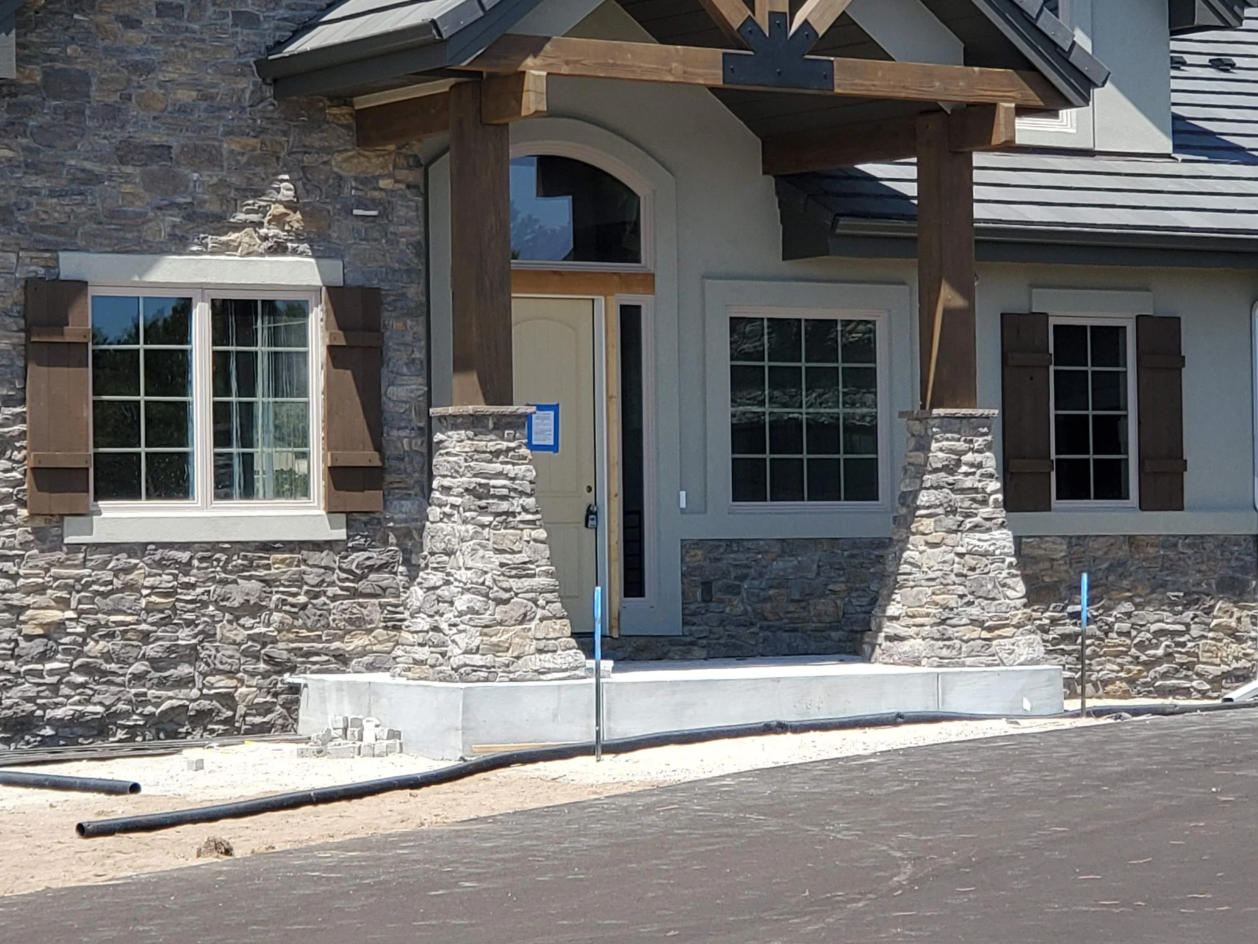 Front of a house under construction with stone and siding exterior, large windows, and a small covered porch with wooden beams.