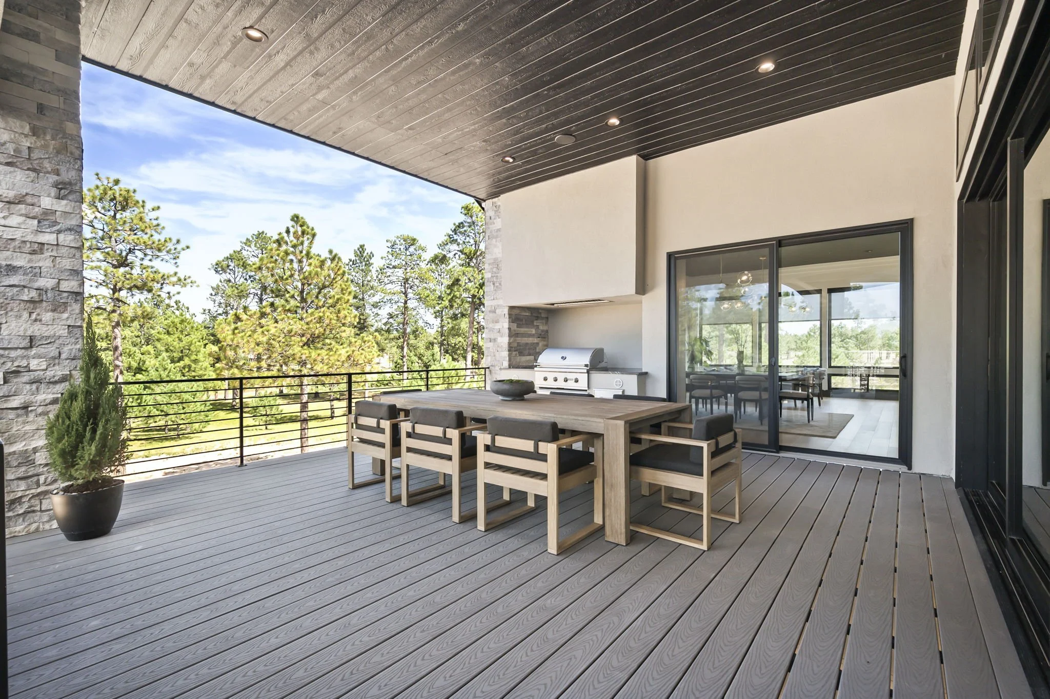 Modern outdoor balcony with wooden dining table, chairs, potted plants, view of trees, sliding glass doors, ceiling with recessed lights.