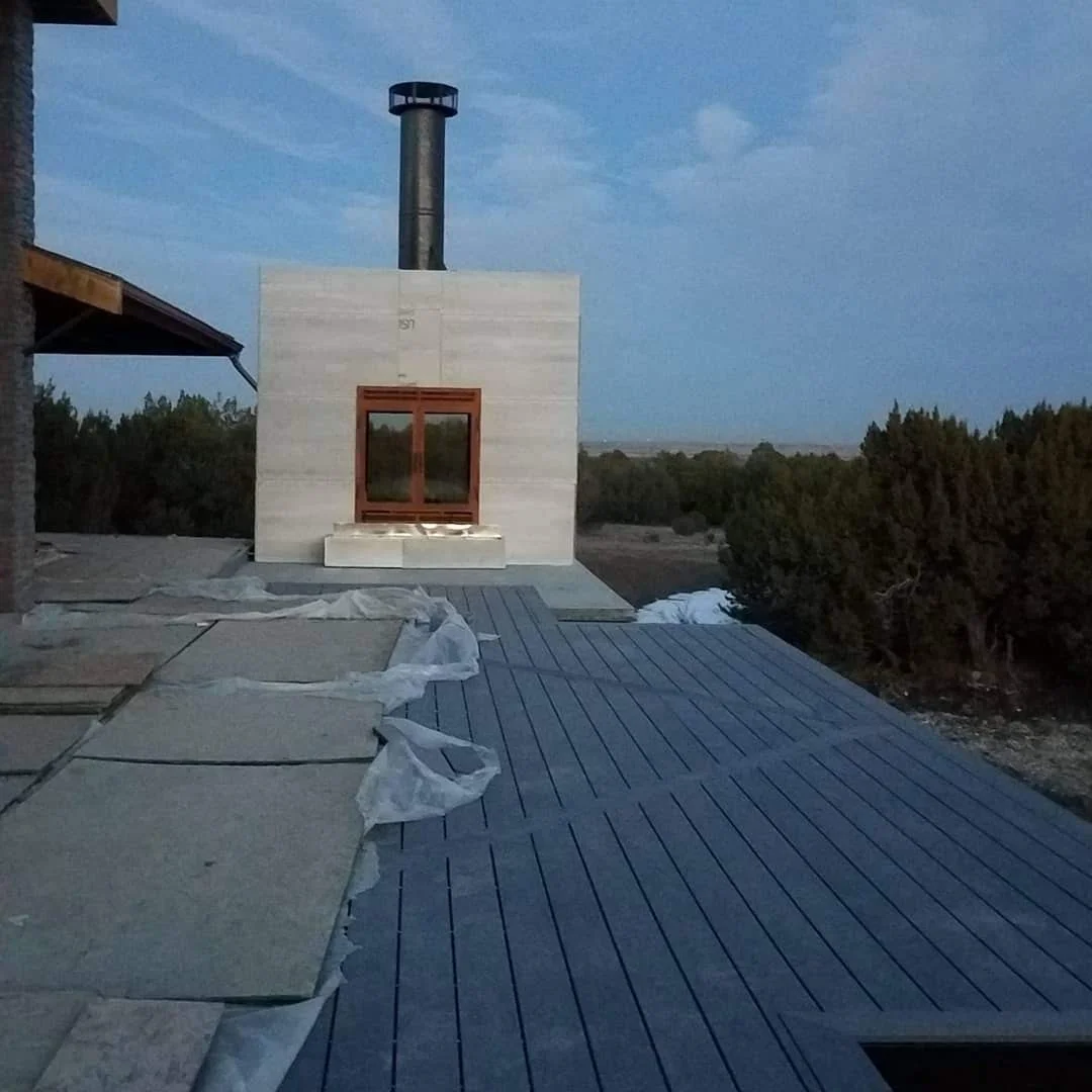 View of a Trex deck with a completed outdoor fireplace in a stone structure, showing a section with slate tiles and another with paving stones, a chimney stack, and surrounding trees with cloudy sky in the background.