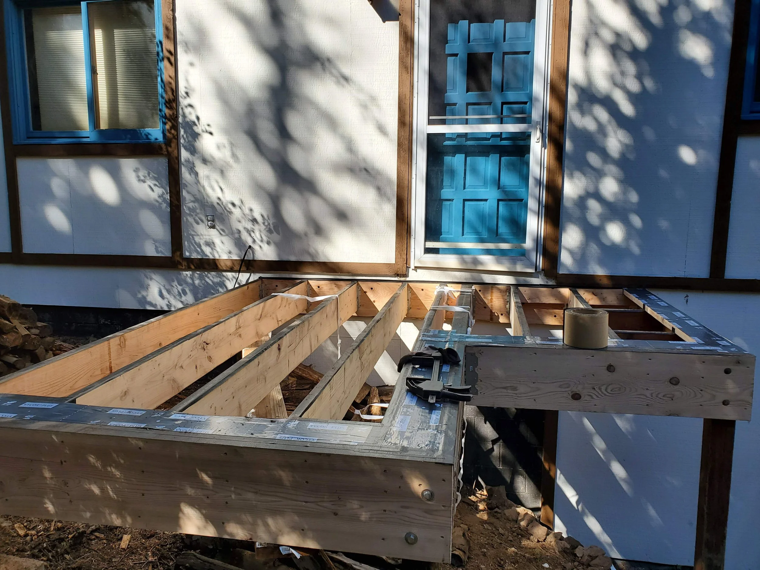 Construction site showing framing for a deck or porch with wooden beams and stairs, next to a house with blue-framed windows and a blue door.