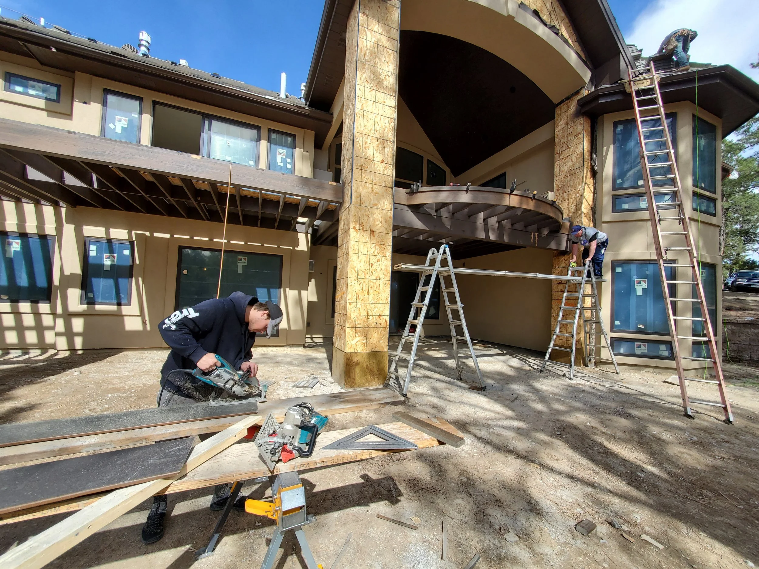 Construction workers are building the exterior of a house, working on the upper level with ladders, tools, and wooden beams, and the house features large windows and stone pillars under a clear blue sky.
