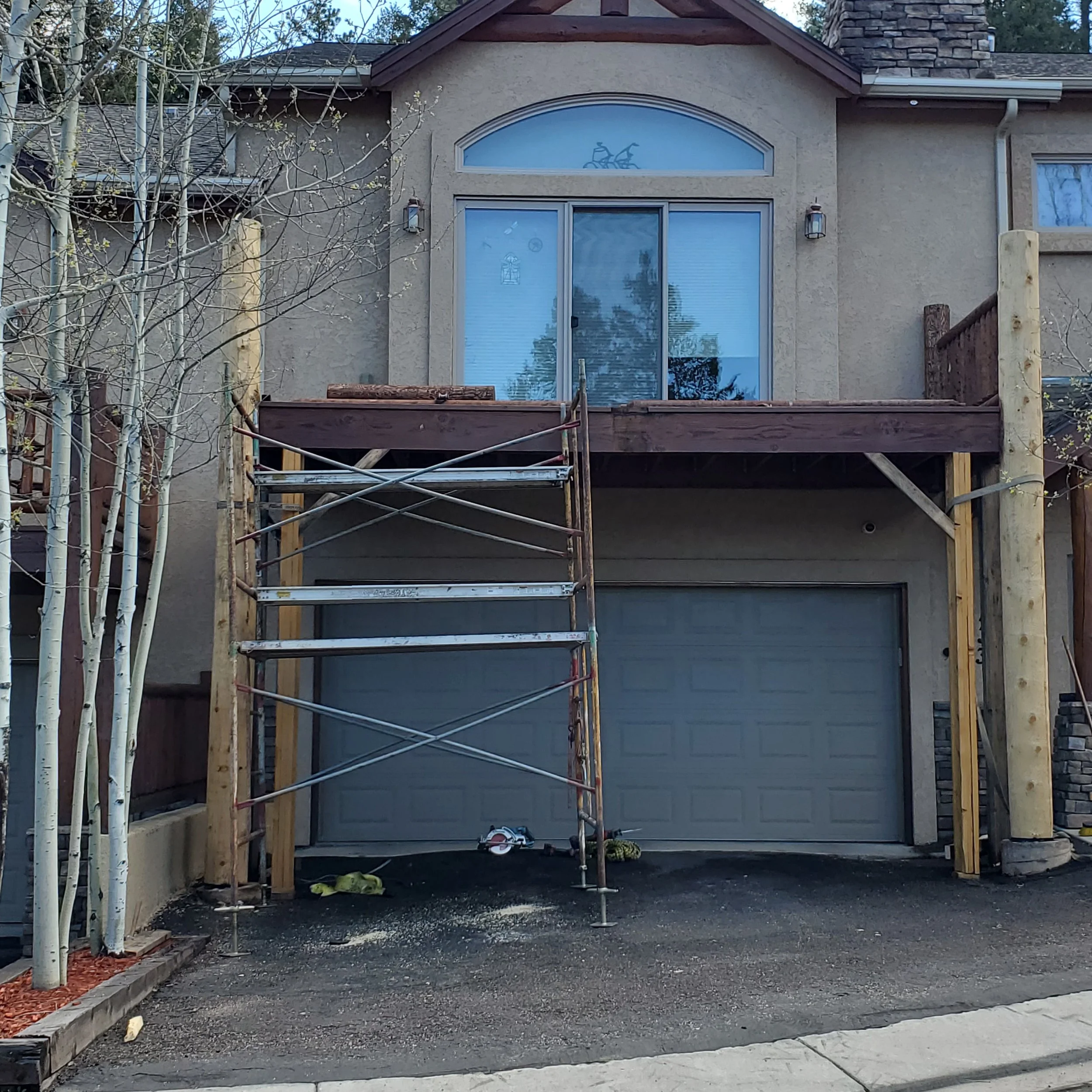 A house under construction with a wooden deck scaffold in front, metal railing, and nearby trees, stone and stucco exterior.