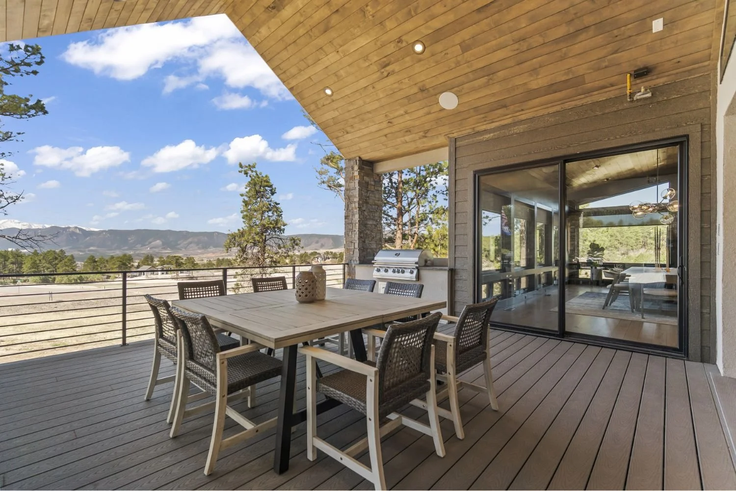 Outdoor patio with wooden flooring and a dining table with six chairs, a barbecue grill, and a railing overlooking a landscape with trees and mountains under a blue sky with clouds.