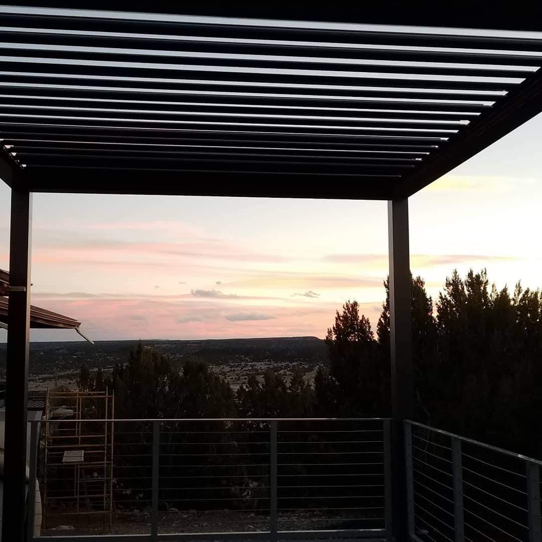 Sunset view from a balcony with a metal pergola and railing, trees in the foreground, and a horizon with pink and orange clouds.