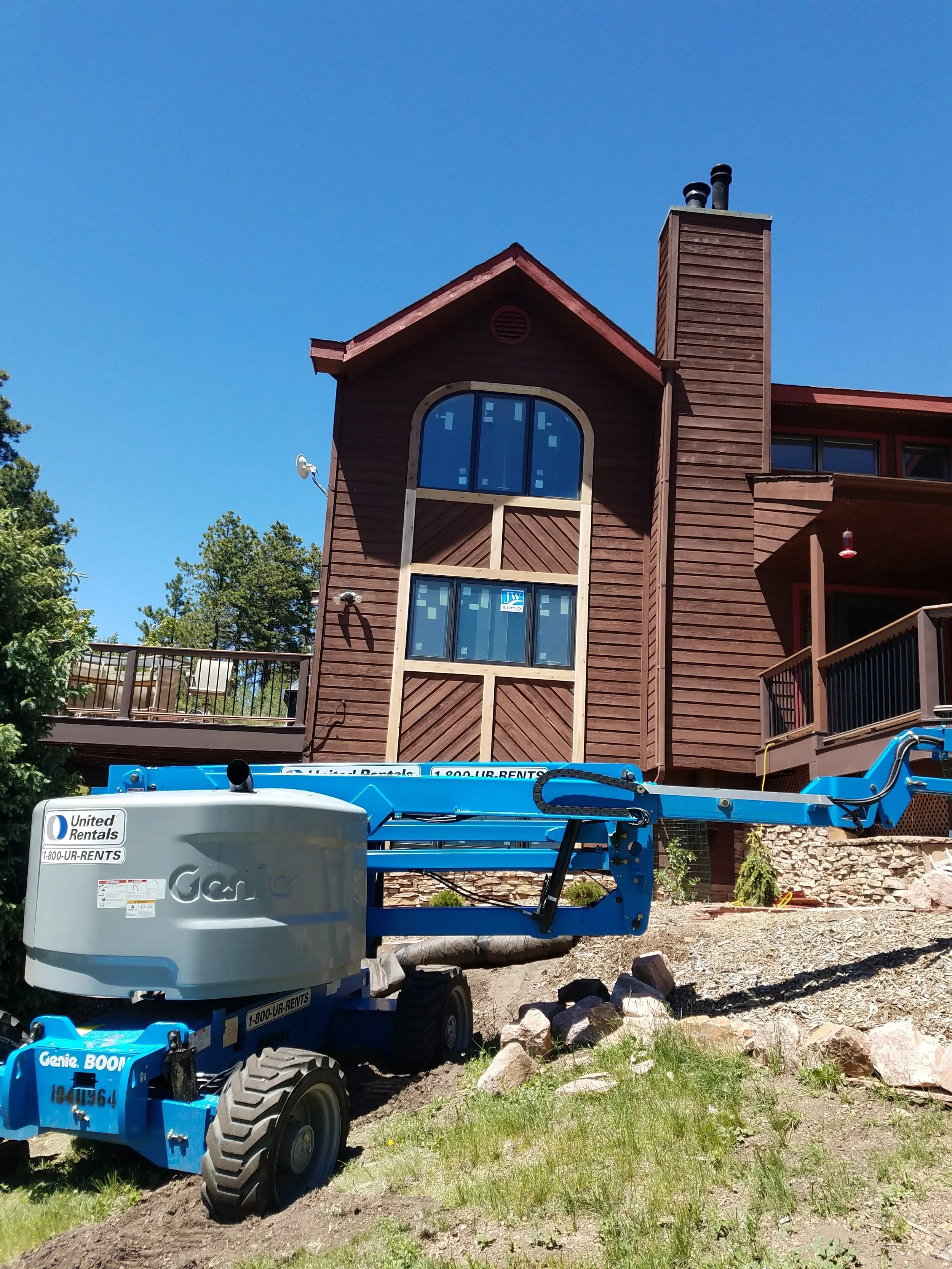 A house with wooden siding and large windows, surrounded by trees and a partly dirt yard, with a blue construction lift in the foreground.