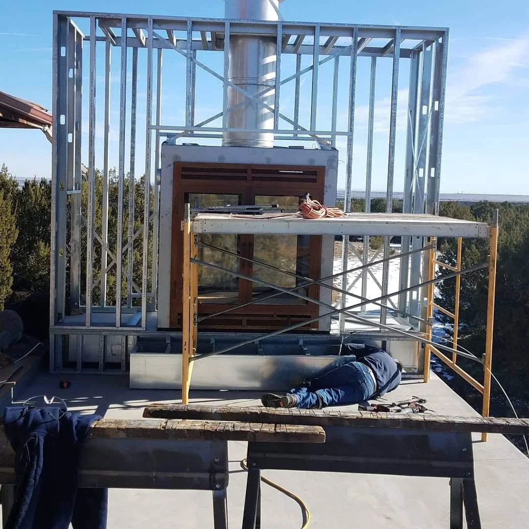 Construction worker working on an outdoor fireplace with metal framing during sunny weather with trees in the background.