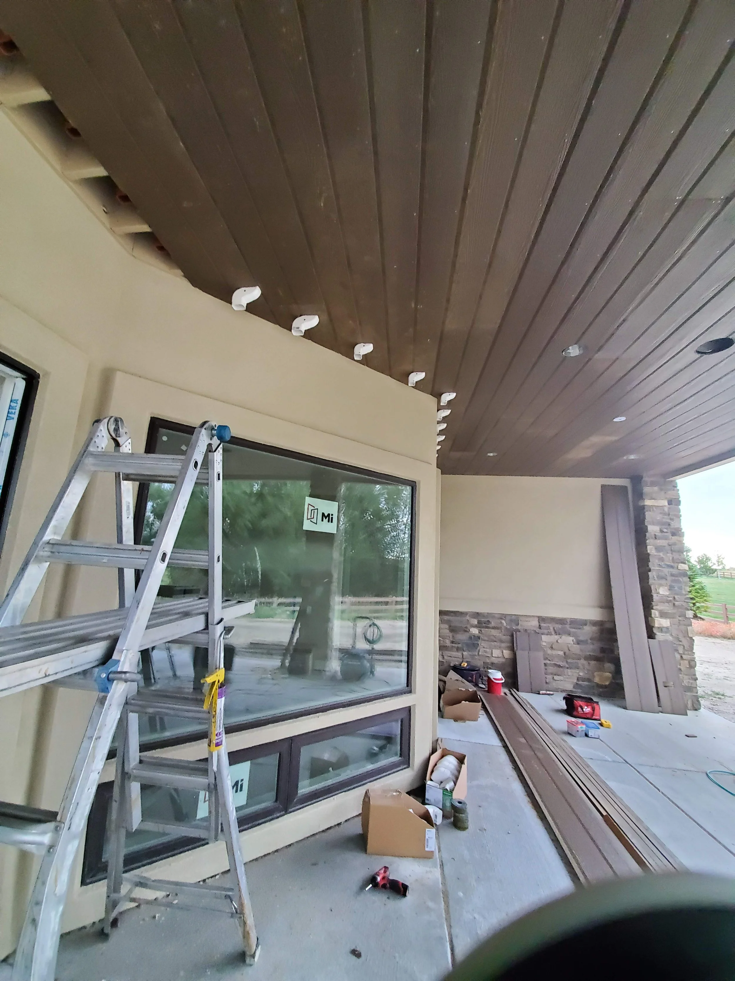 View of a room under construction or renovation, with a ladder, tools, construction materials, and a large window, featuring a wooden ceiling and brick accents.