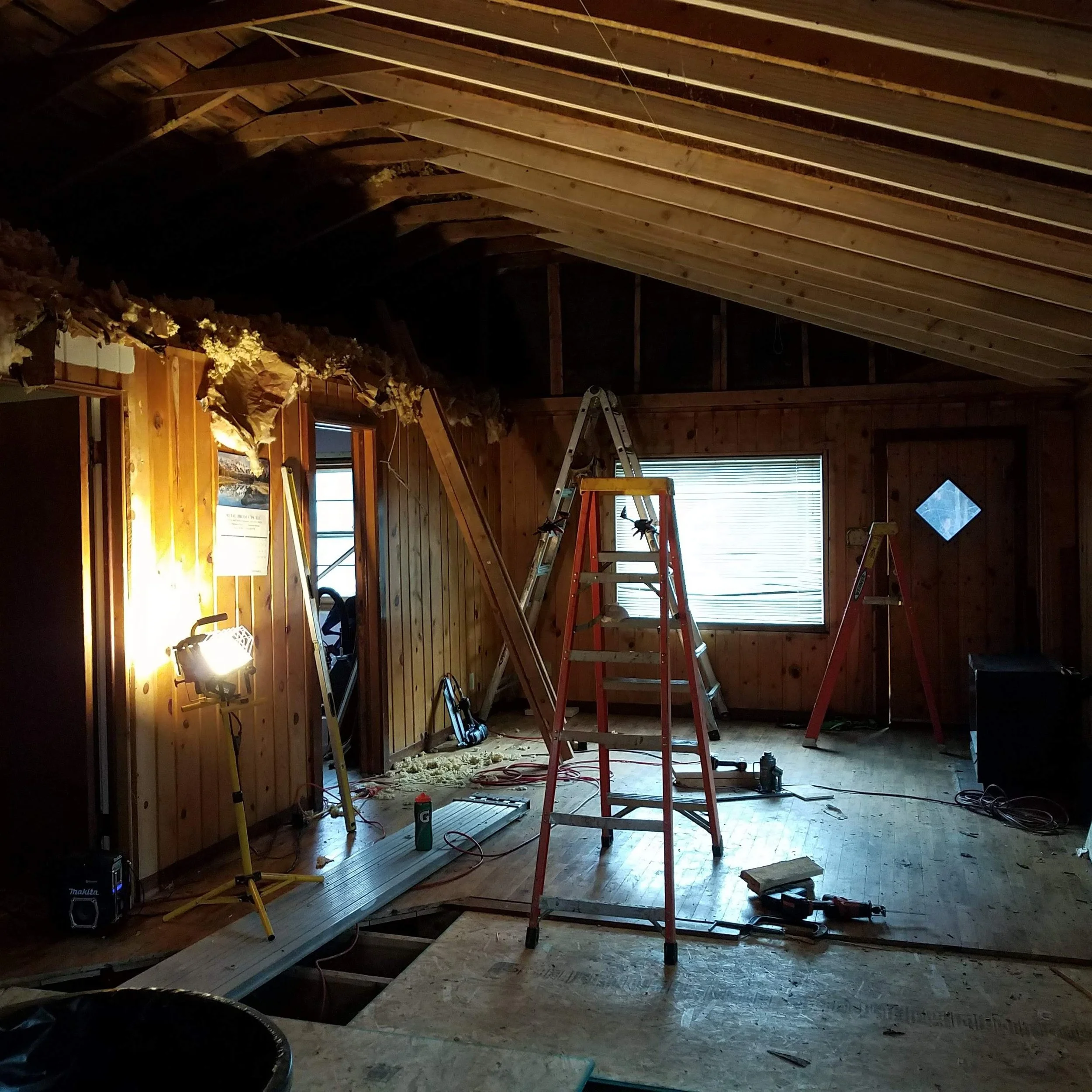 The image shows an attic under construction or renovation. It features exposed wooden beams on the ceiling, a step ladder, and construction tools scattered around. Insulation material is visible in the upper left corner, and there is a window with bl