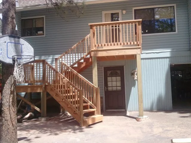 Backyard of a house with a new wooden deck and staircase leading to an upper level, with a basketball hoop attached to a tree on the left.