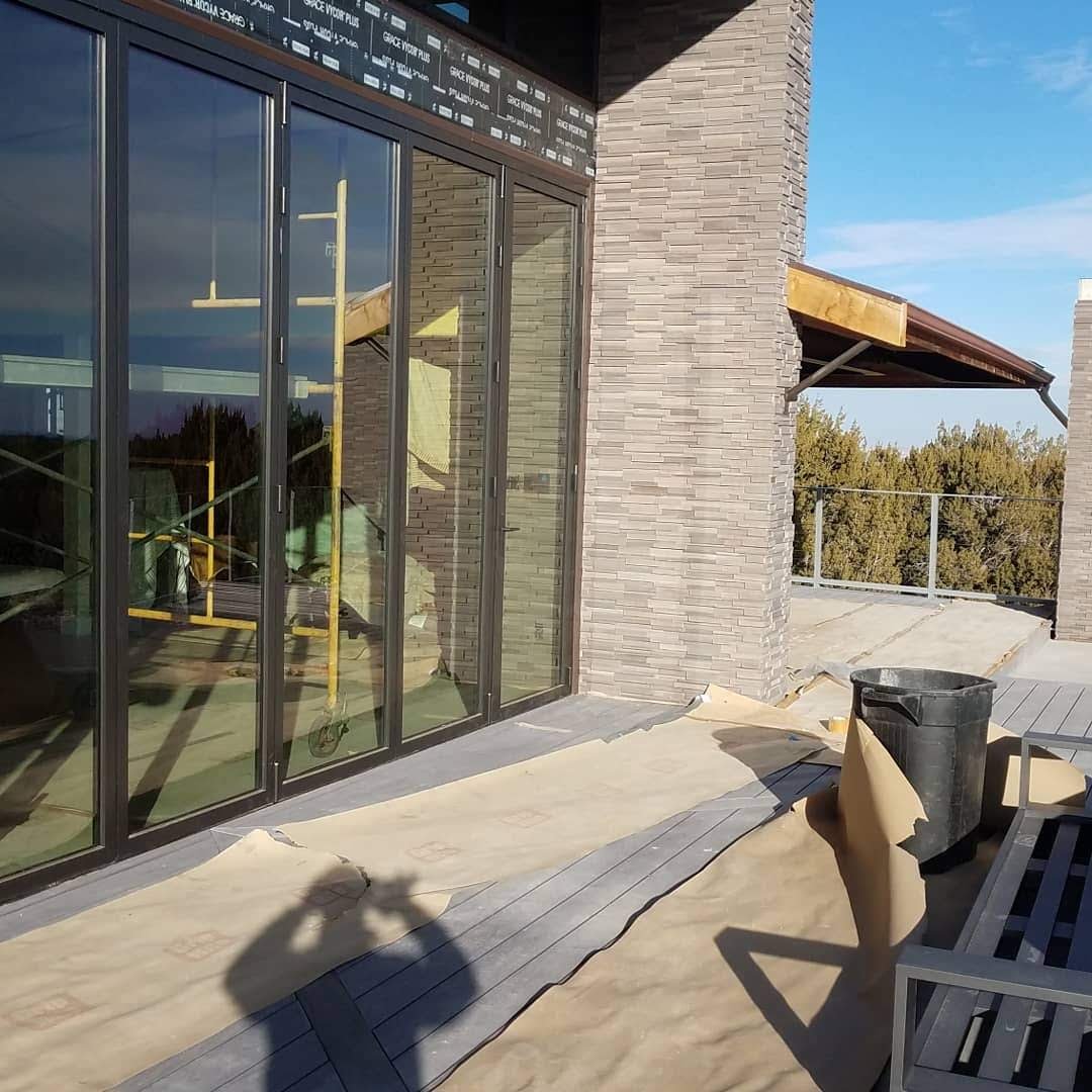 Building exterior with large glass sliding doors, brick wall, wooden deck, and construction materials including paper, a trash can, and a bench on a sunny day.