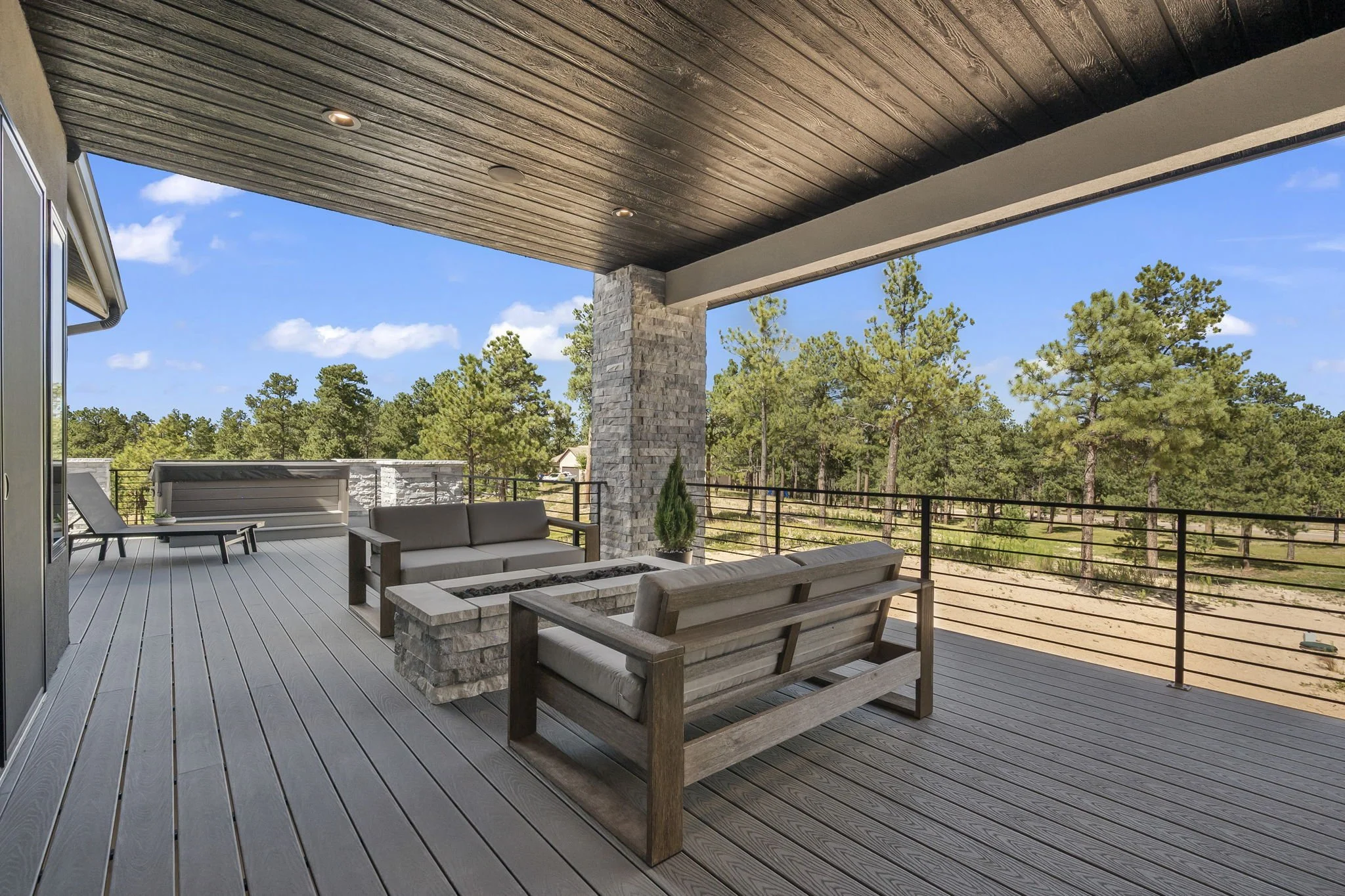 Covered outdoor patio with gray wooden flooring, wooden seating, gray cushioned chairs, fire pit, and a view of trees and blue sky.