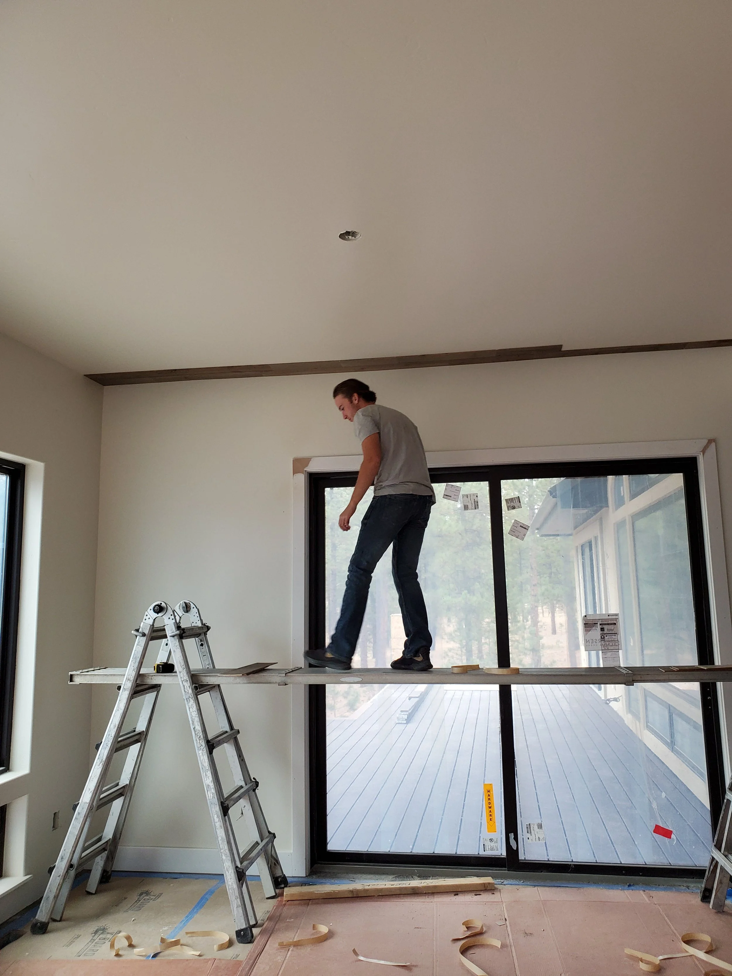 A man standing on a ladder inside a room, installing a large glass door or window, with construction materials and tools around him.