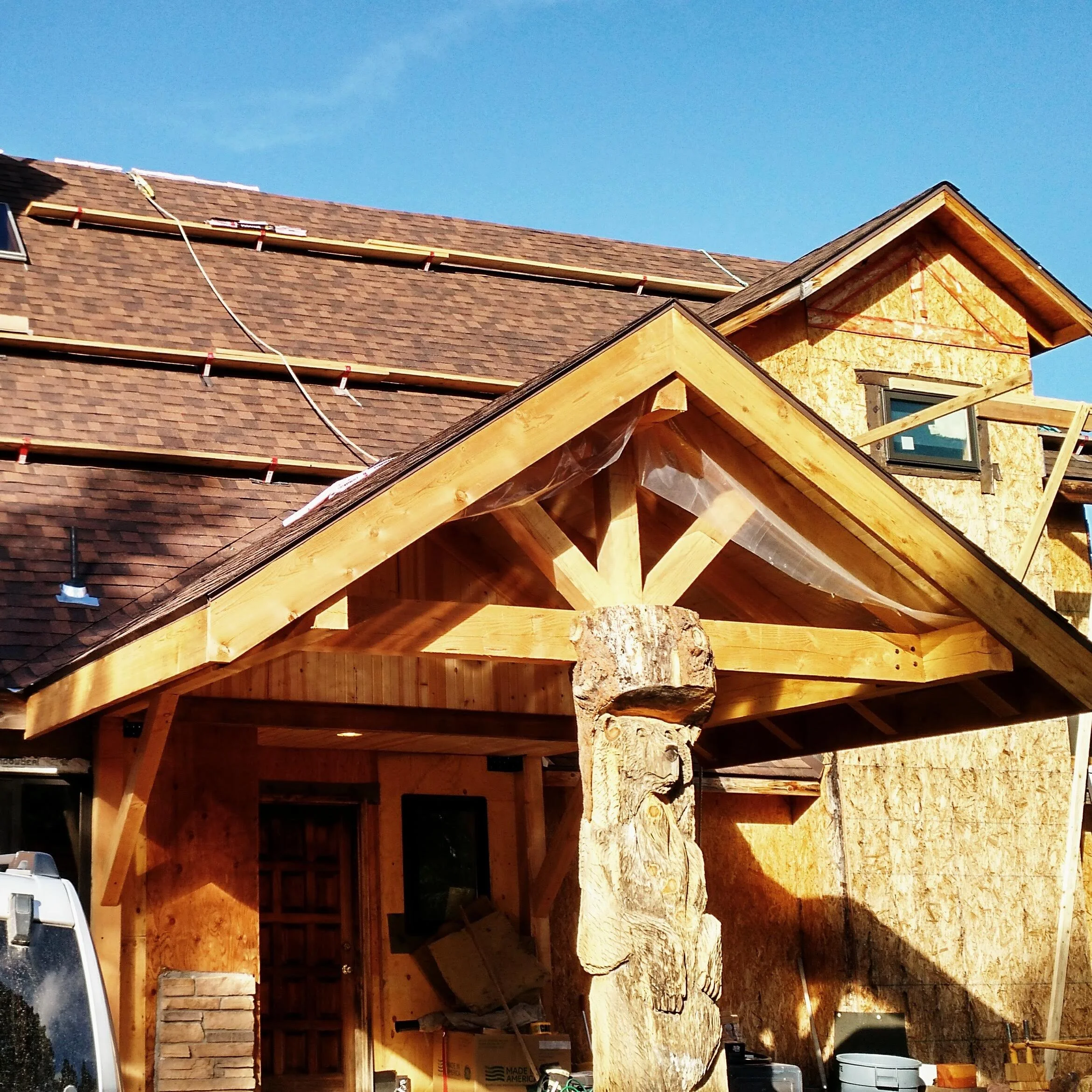 Construction site of a house with a wooden frame, a partially built roof with new shingles, and ongoing roofing work under a clear blue sky.
