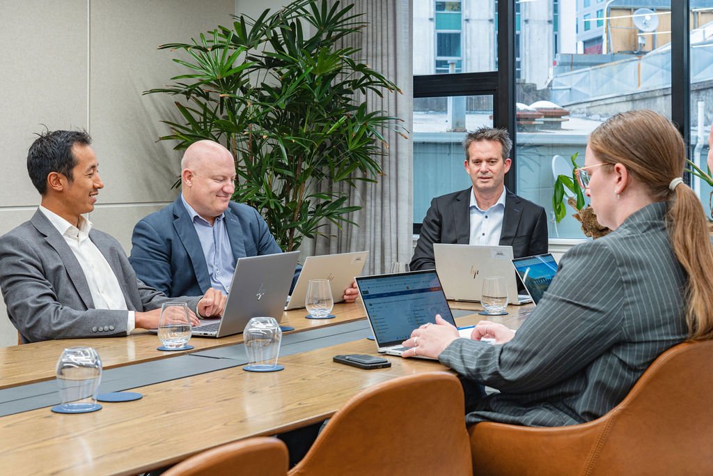 Four professionals in business attire seated at a conference table with laptops and glasses of water, engaging in a meeting in a well-lit office with large windows and indoor plants.